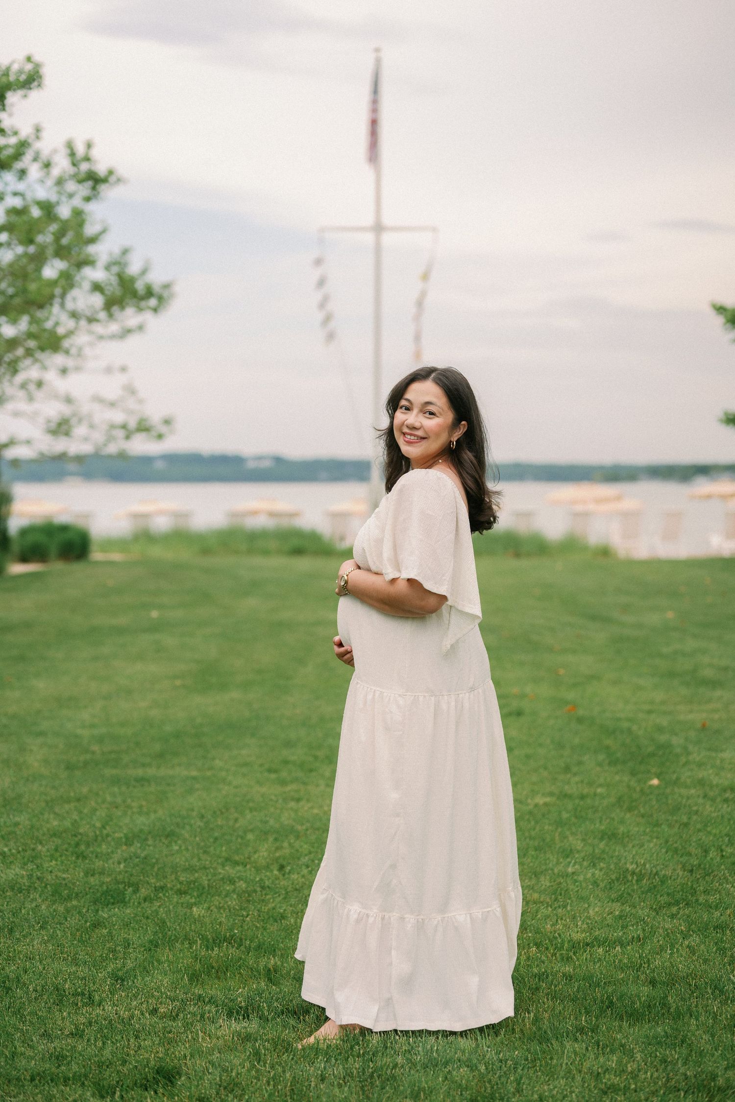 Smiling pregnant woman in white maxi dress standing on green lawn near waterfront at dusk.
