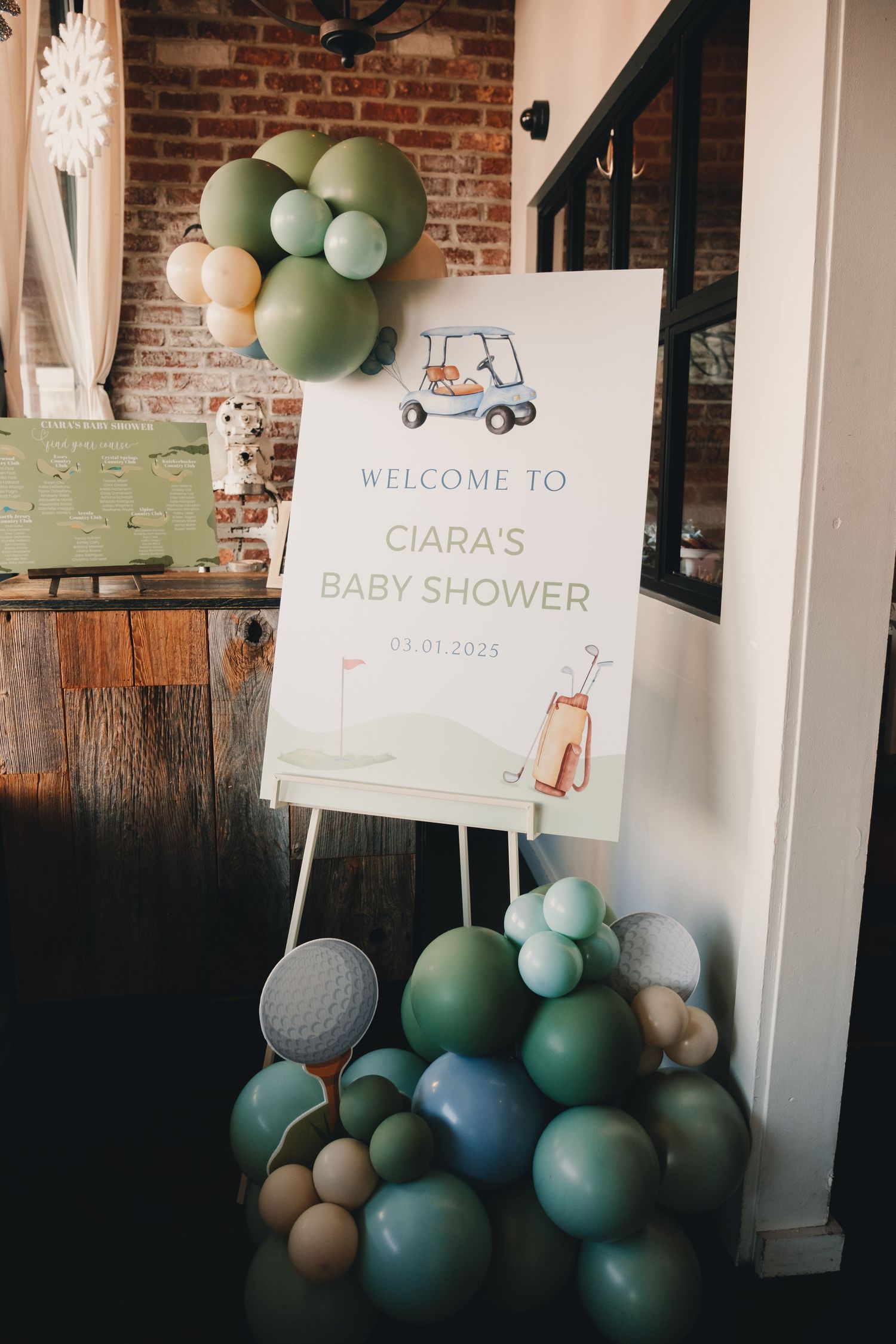 Baby shower decorations featuring mint green and white balloons against an exposed brick wall with welcome signage.