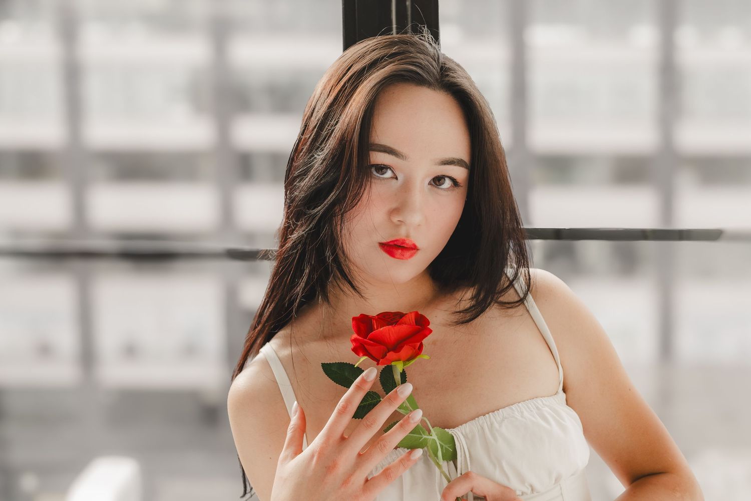 Young woman with long dark hair and red lipstick holding a red rose against her chest, wearing a white sleeveless top by a bright window.