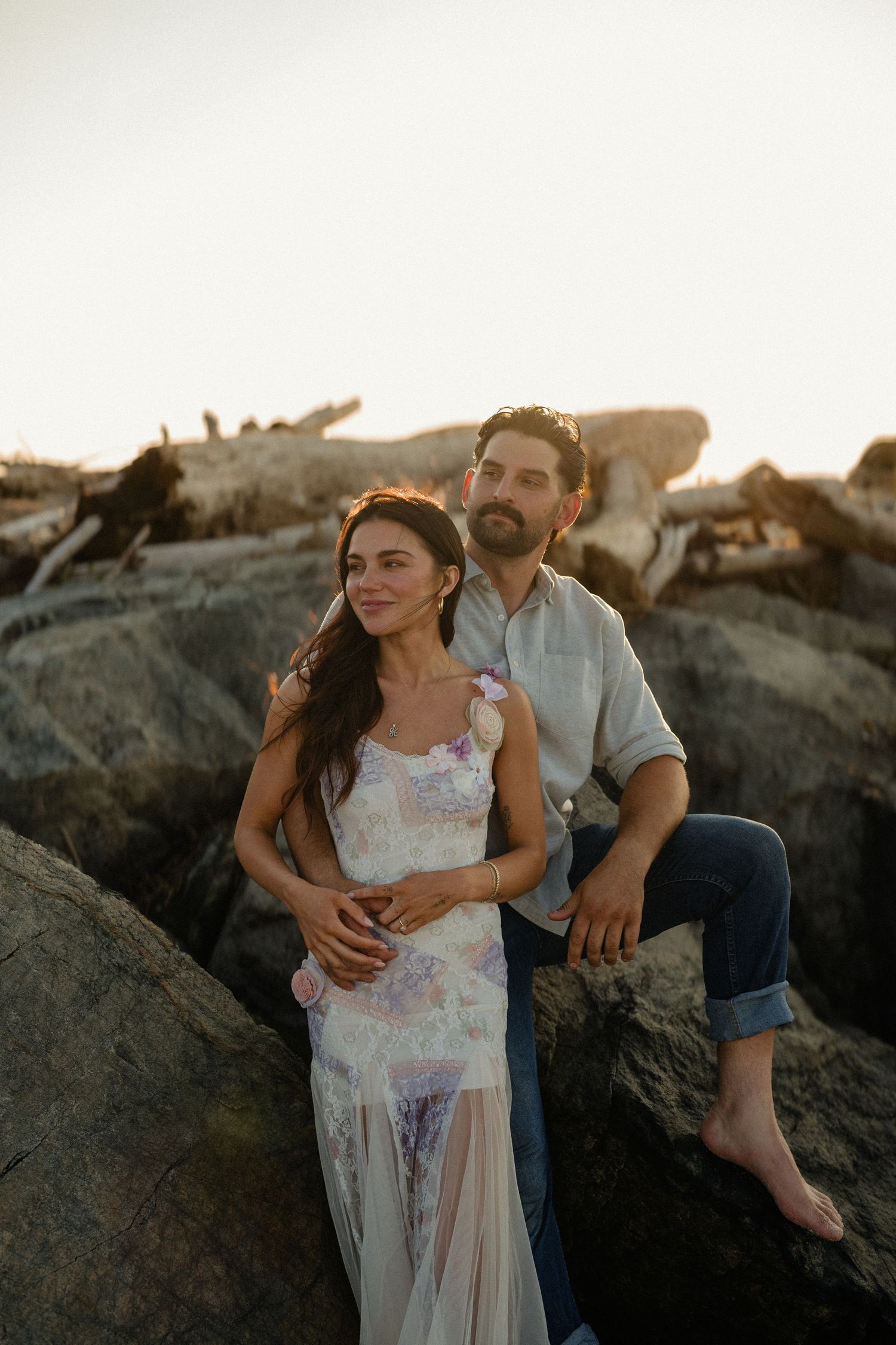 A couple sits on coastal rocks at sunset, woman in floral dress, man in casual attire, with driftwood backdrop.