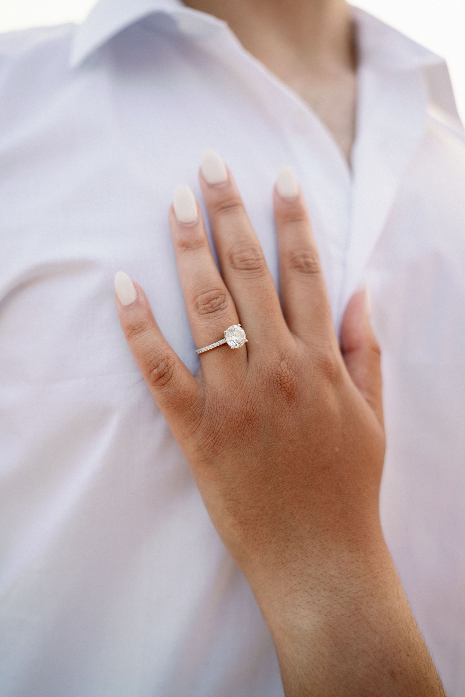 Close-up of a hand with nude nails wearing an elegant oval diamond solitaire engagement ring on a white shirt background.