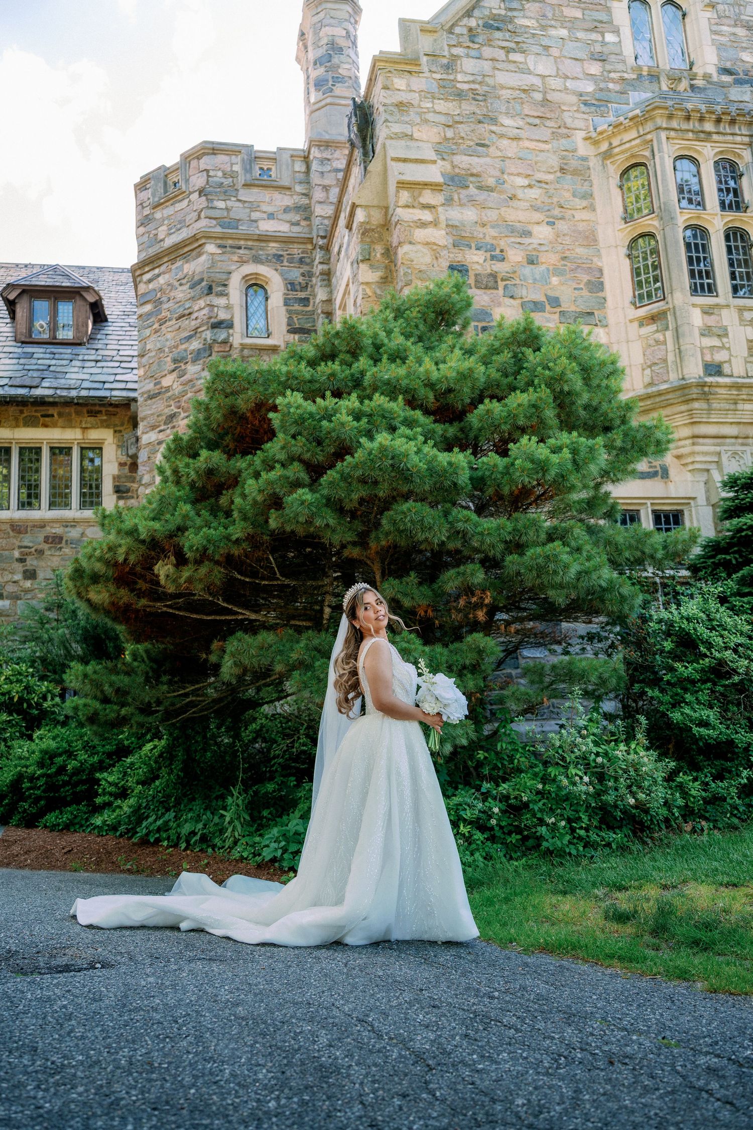 Bride in white gown with long train holds bouquet outside stone castle building with lush green trees.