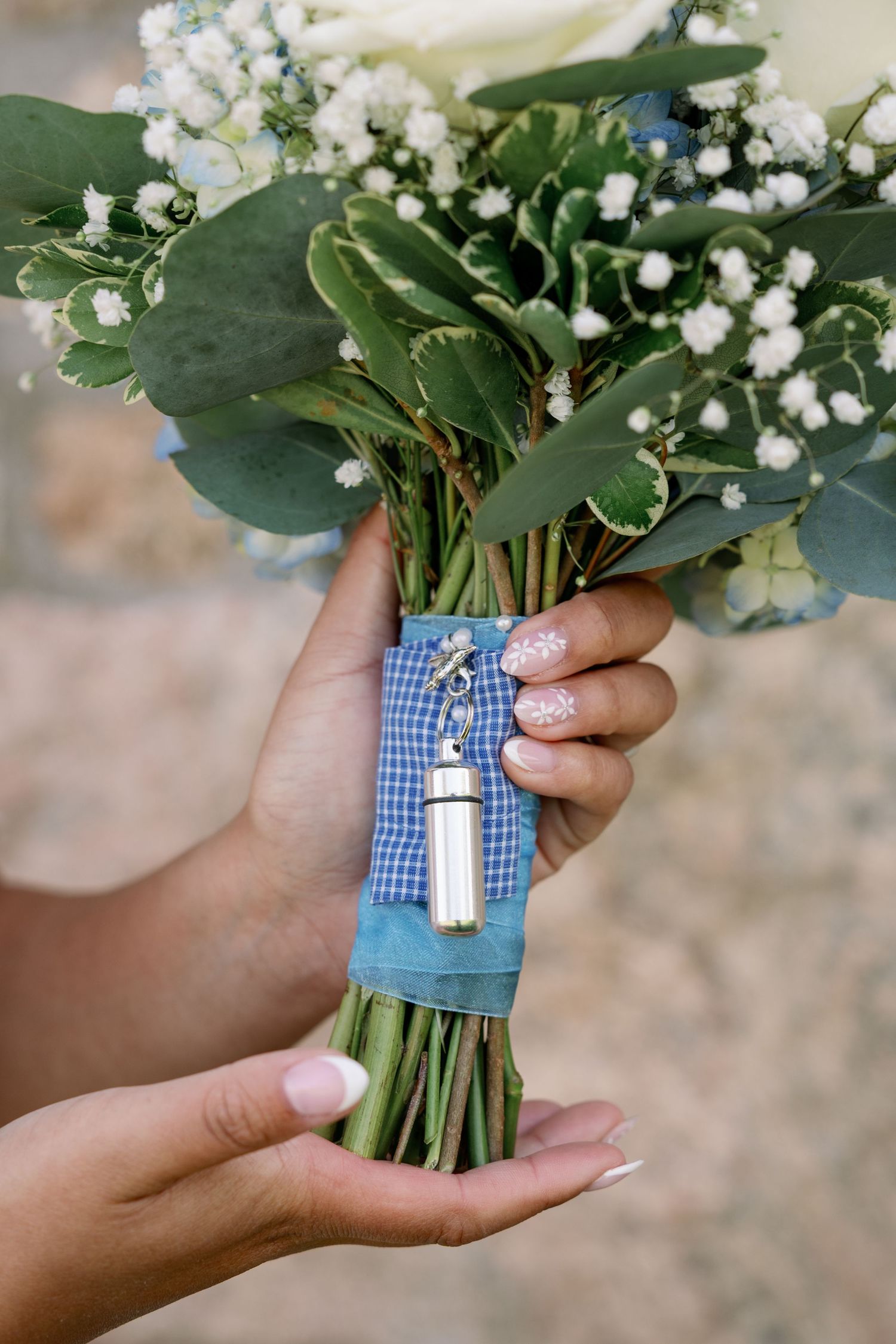 Hands holding a bridal bouquet of baby's breath and green leaves wrapped in blue gingham ribbon with a silver memorial charm.