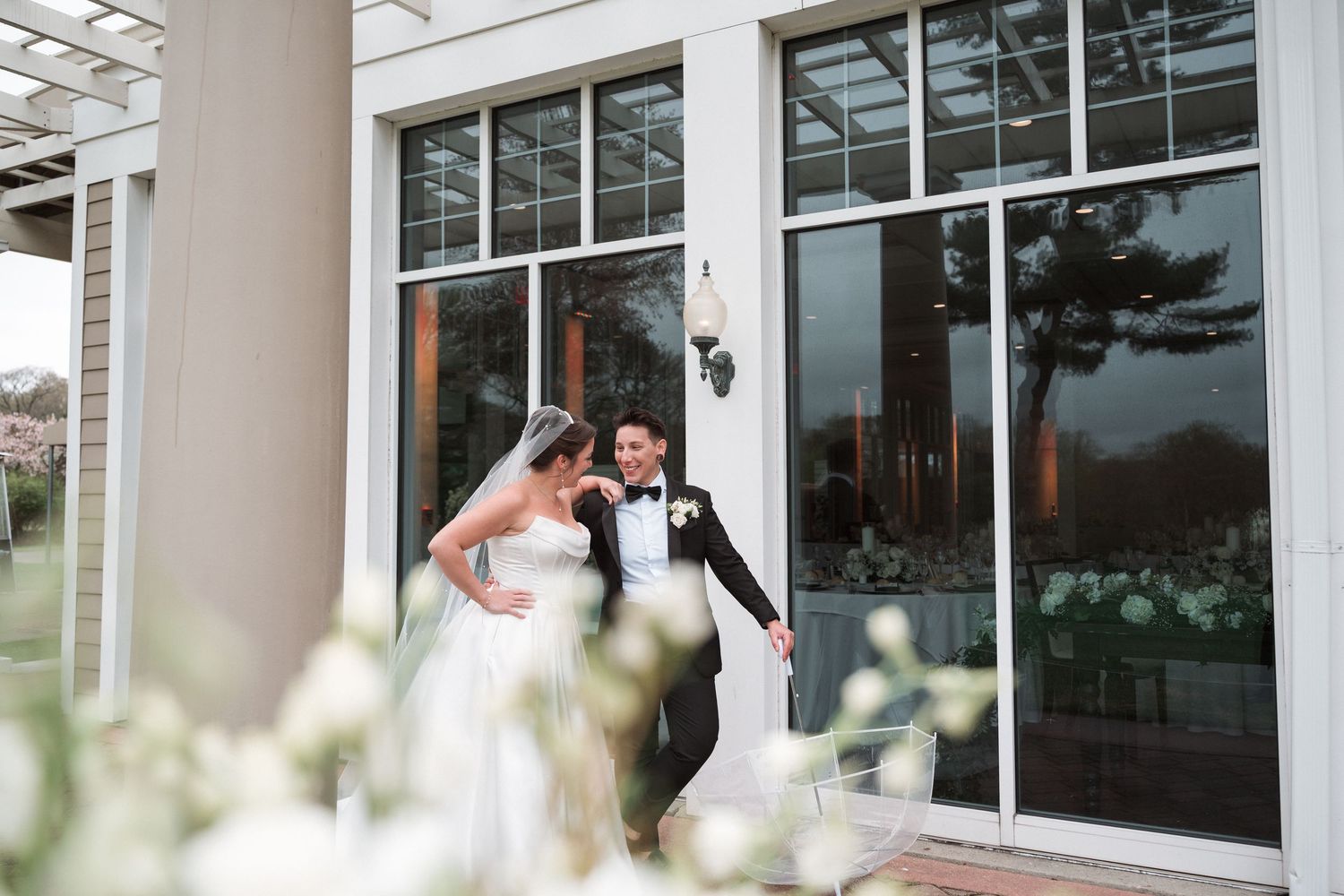 Bride and groom share a romantic moment outside elegant venue with white floral details and large glass windows.
