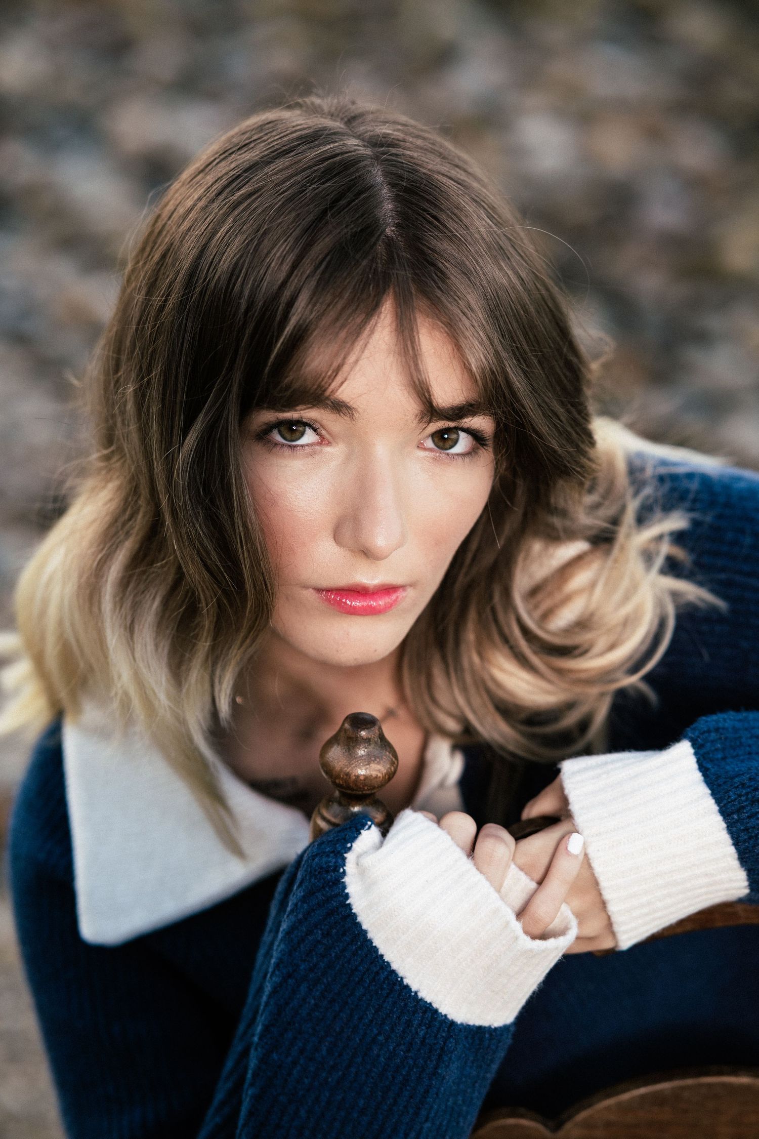 Young woman with wavy brown ombre hair and bangs gazes at the camera in a navy knit sweater with white collar and cuffs, resting on a chair.