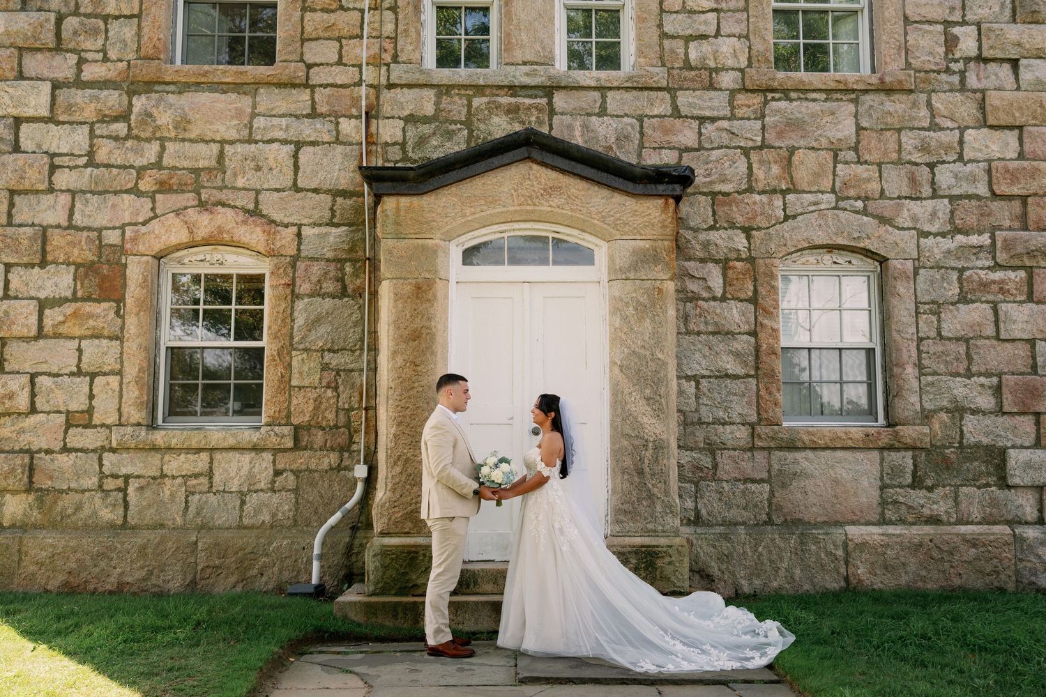 Bride in a long white gown and groom in a light suit hold hands in front of a white door on a historic stone building.