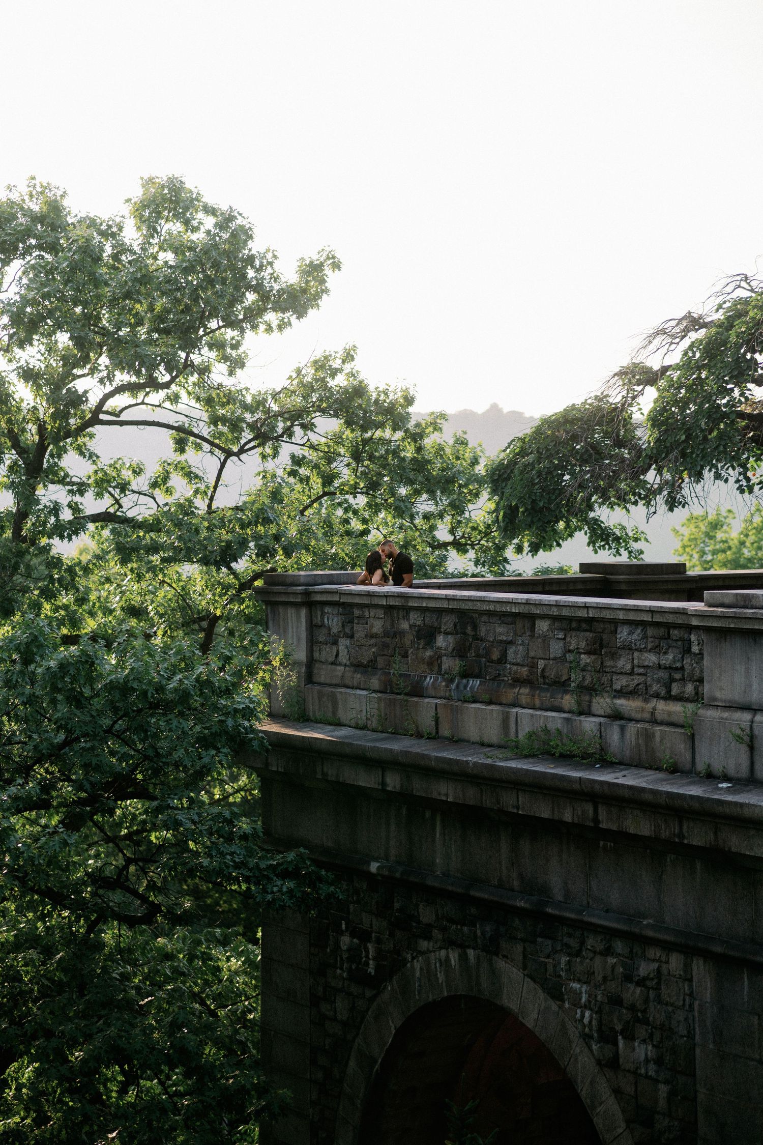 Two people stand atop a historic stone bridge arch surrounded by lush green trees in a serene park setting.