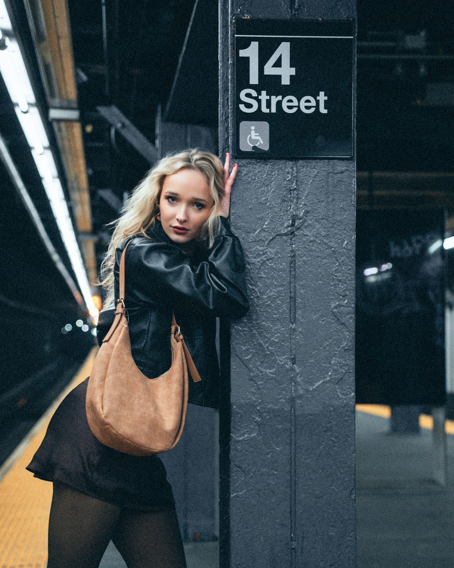 Woman with blonde hair in a black leather jacket leans on a pillar by a 14 Street subway sign, holding a tan suede shoulder bag.