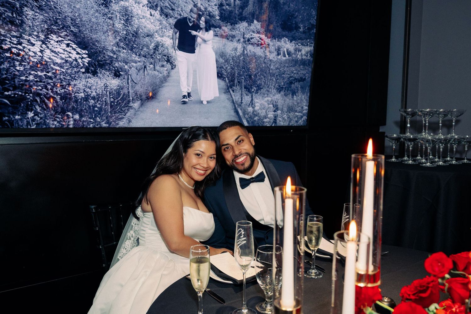Bride and groom smiling at wedding reception table with champagne, candles, and red roses, with wedding photo projected behind them.