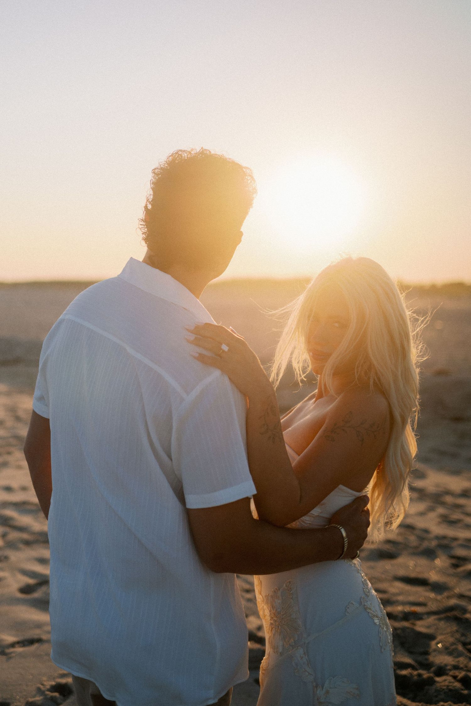Couple embracing on a sandy beach at golden sunset, woman with blonde hair in white dress, romantic outdoor photography.