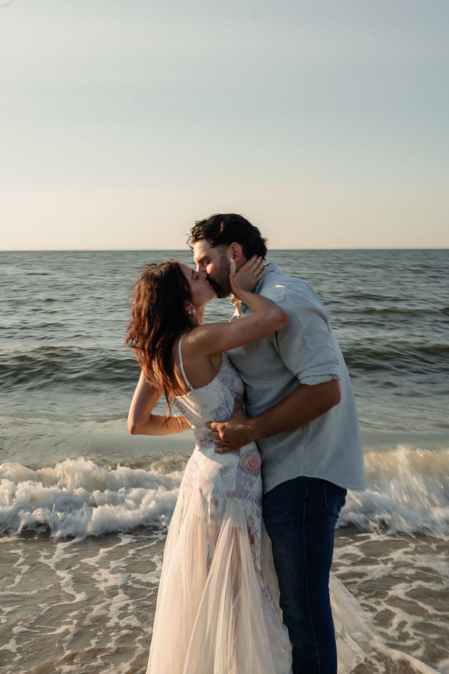 Couple sharing a romantic kiss on a sandy beach at sunset, waves crashing behind them.