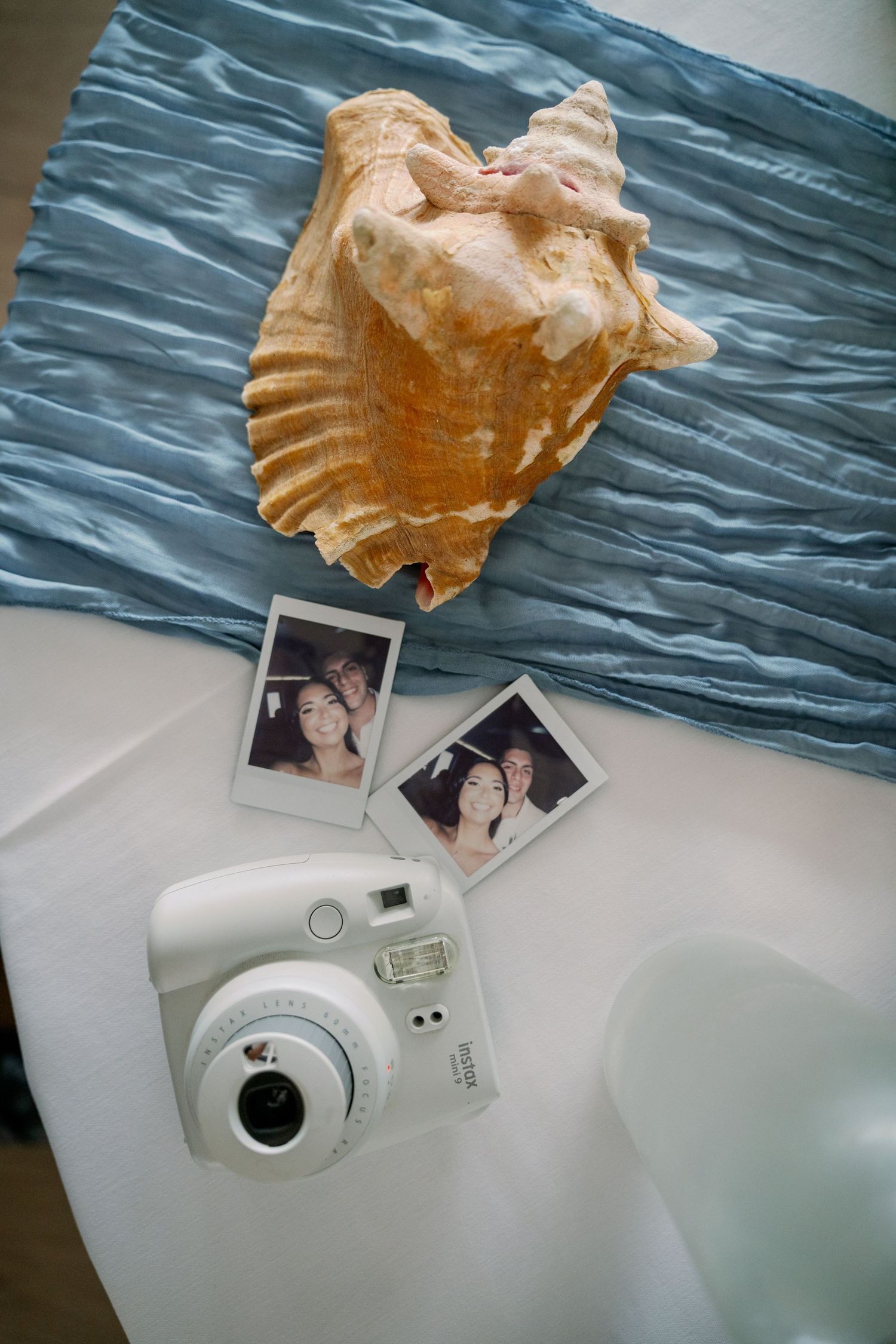 Large conch shell on blue crinkled fabric beside a white Instax instant camera and two Polaroid-style photos of a smiling couple.