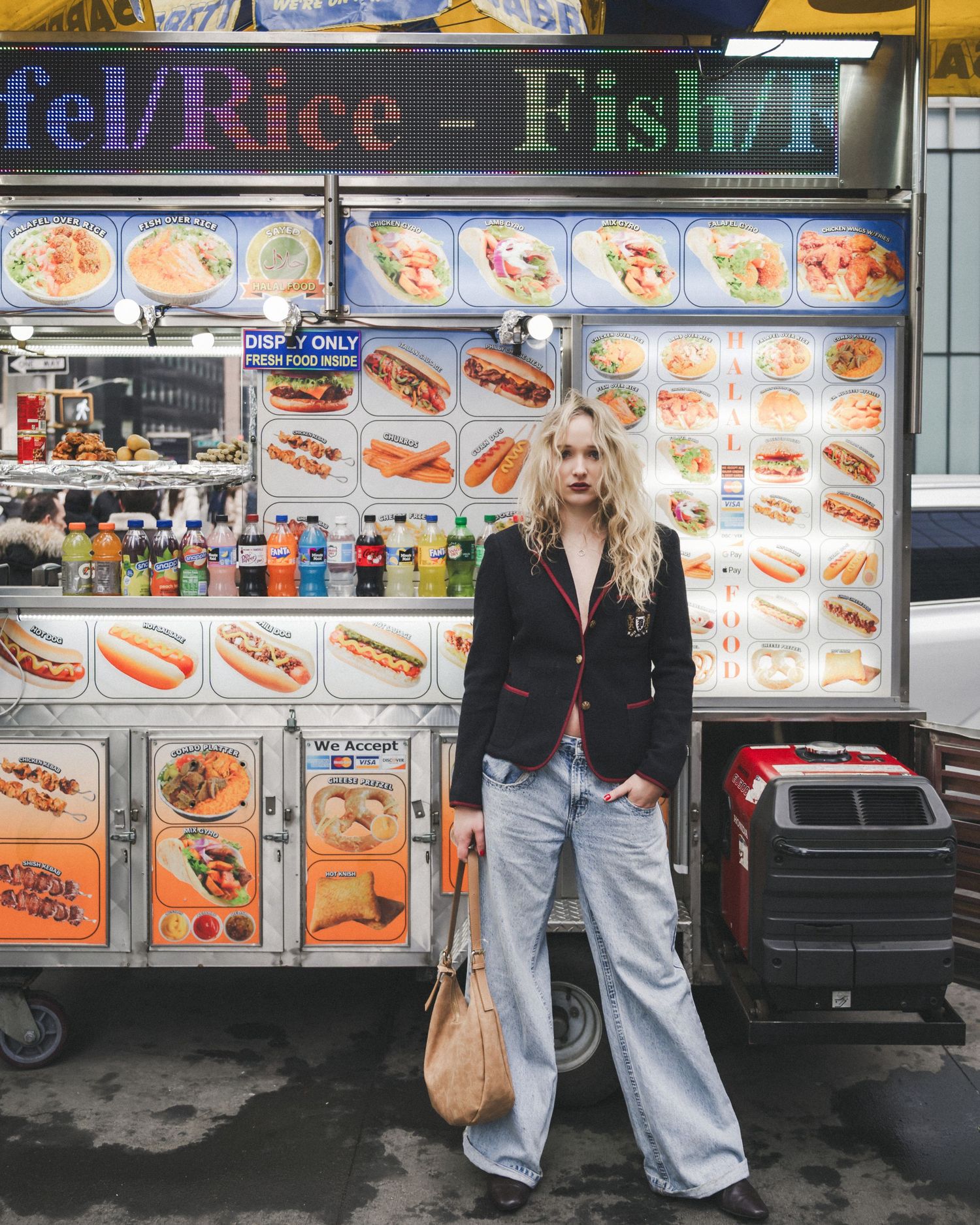 Woman with wavy blonde hair in a navy blazer and wide-leg jeans holds a tan bag in front of a colorful New York street food cart.