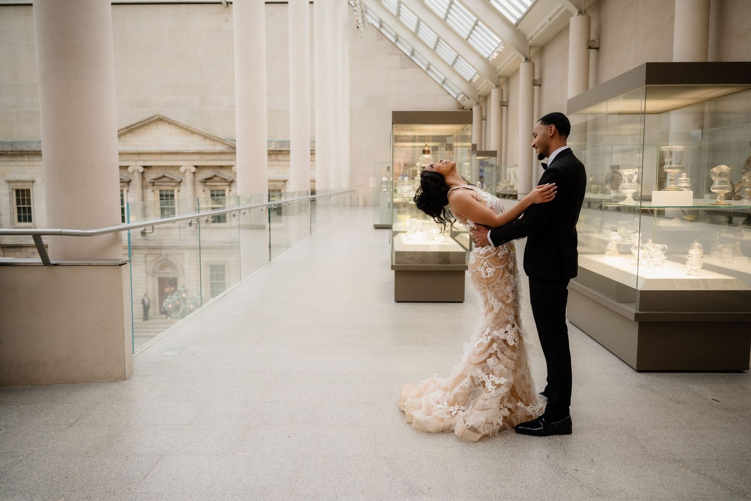 A couple in wedding attire embraces in an elegant museum gallery with marble columns and display cases.