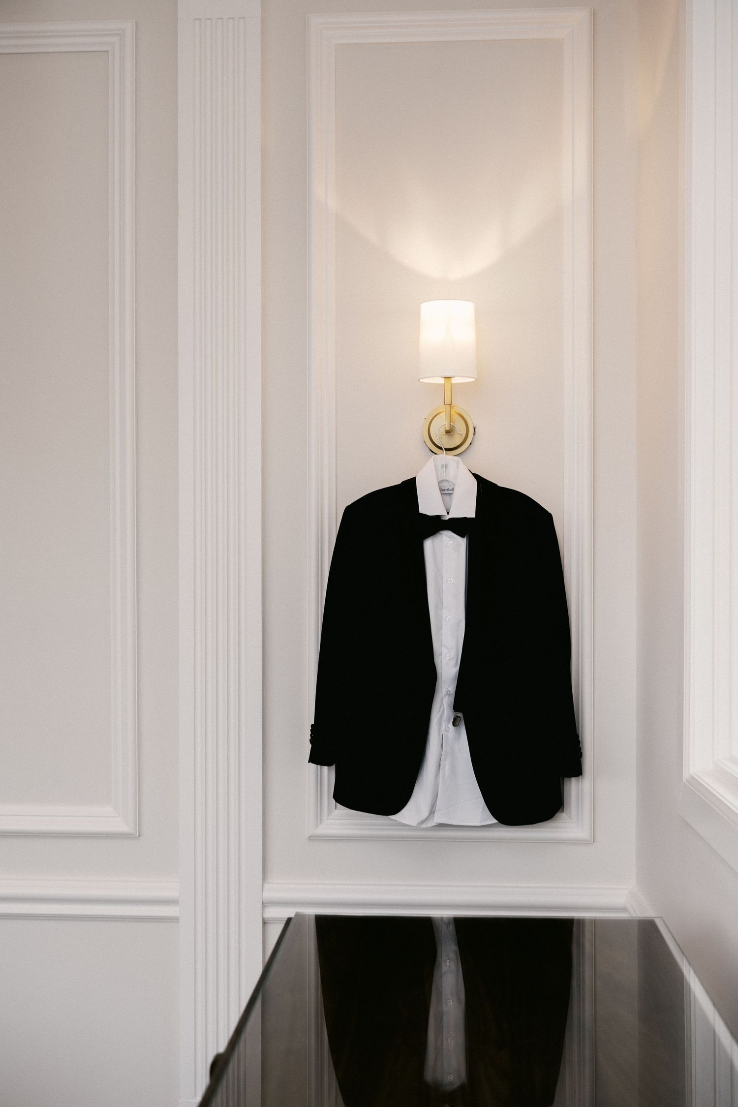 Black tuxedo jacket with white shirt and bow tie hanging on wall hook beneath elegant gold sconce light.