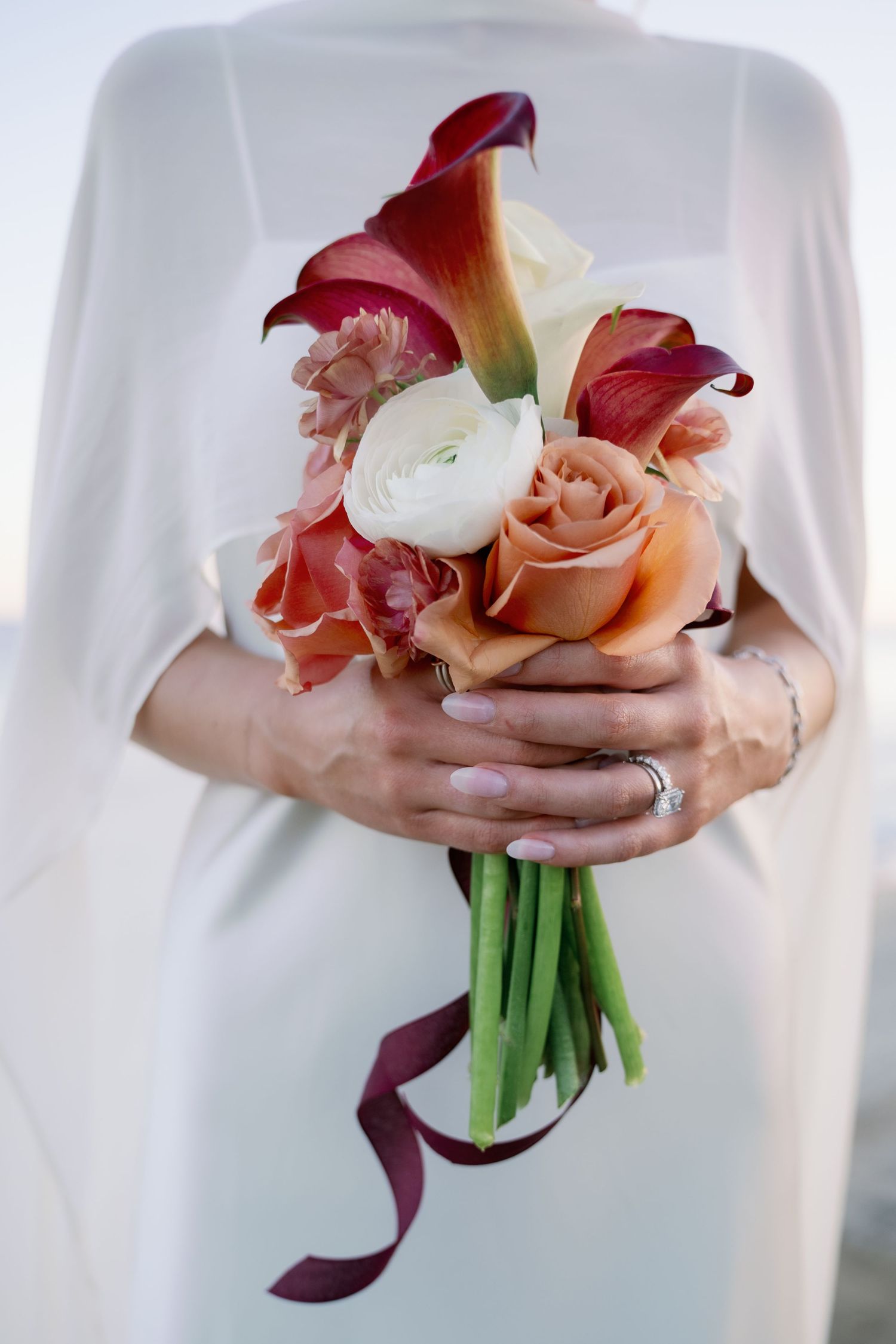 Bride in a white cape dress holds a small bouquet of peach roses, white ranunculus and burgundy calla lilies tied with a satin ribbon.