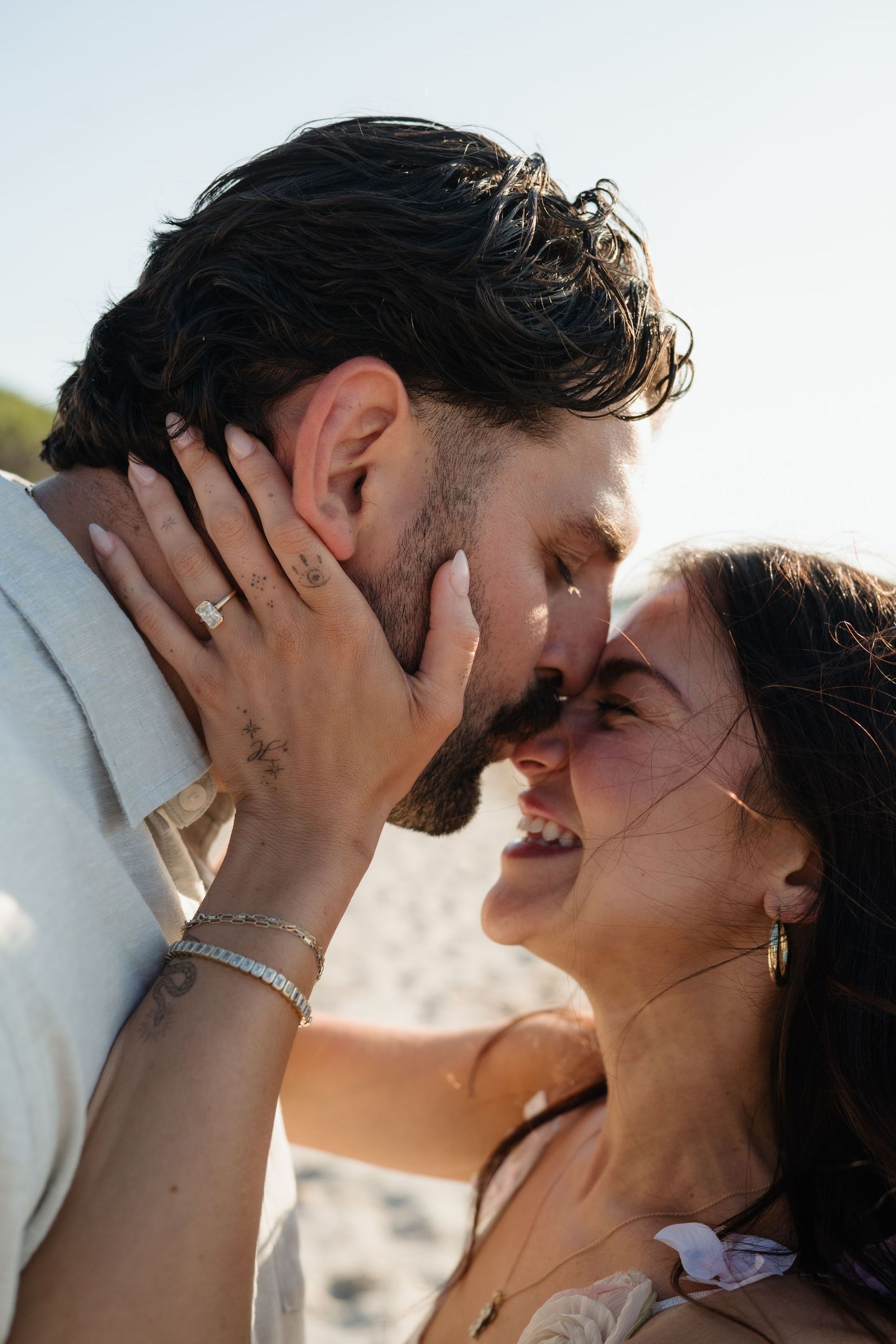A couple shares a romantic almost-kiss on a sunny beach, woman smiling with hand on mans face, wearing a ring and bracelet.
