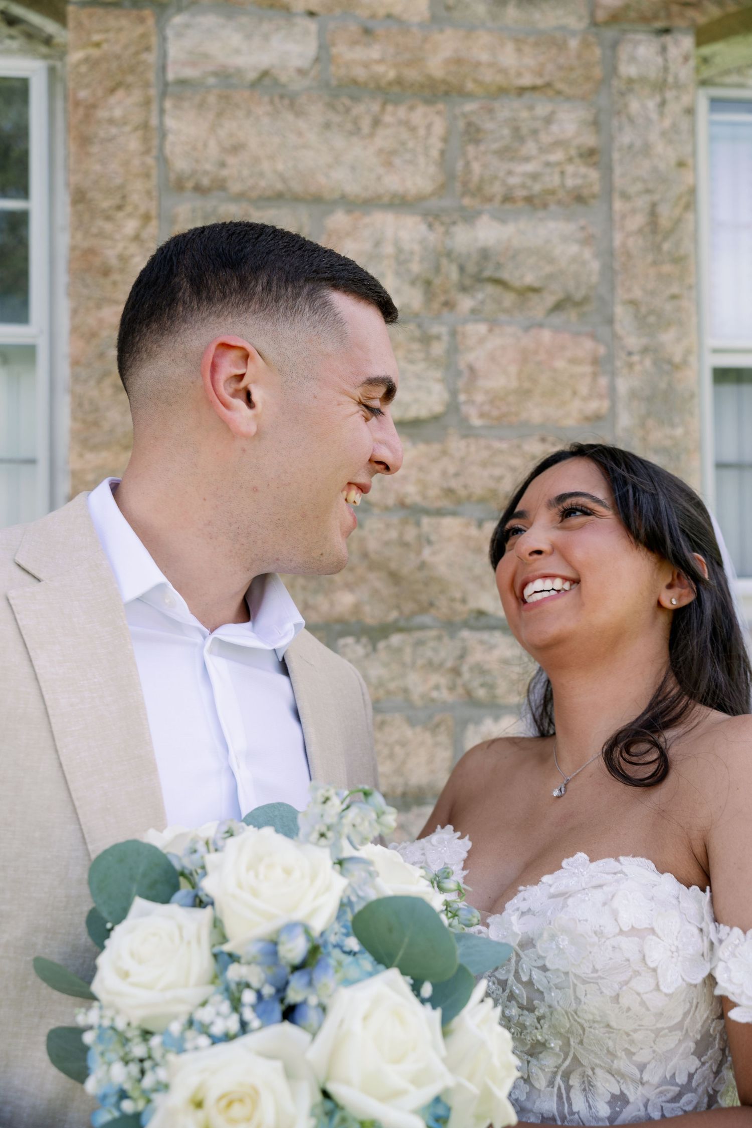 Smiling bride in a white floral lace gown and groom in a tan suit share a joyful moment beside a bouquet of white roses.