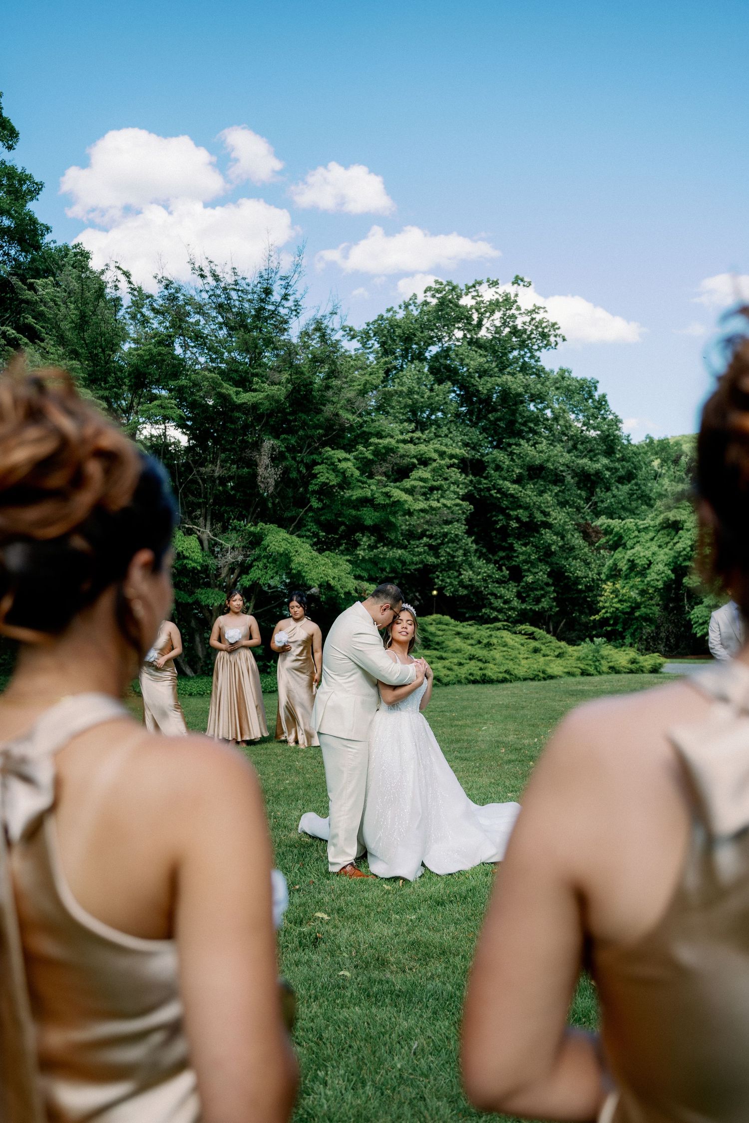 Bride and groom embrace during outdoor wedding ceremony on lush green lawn, surrounded by bridesmaids in gold dresses.