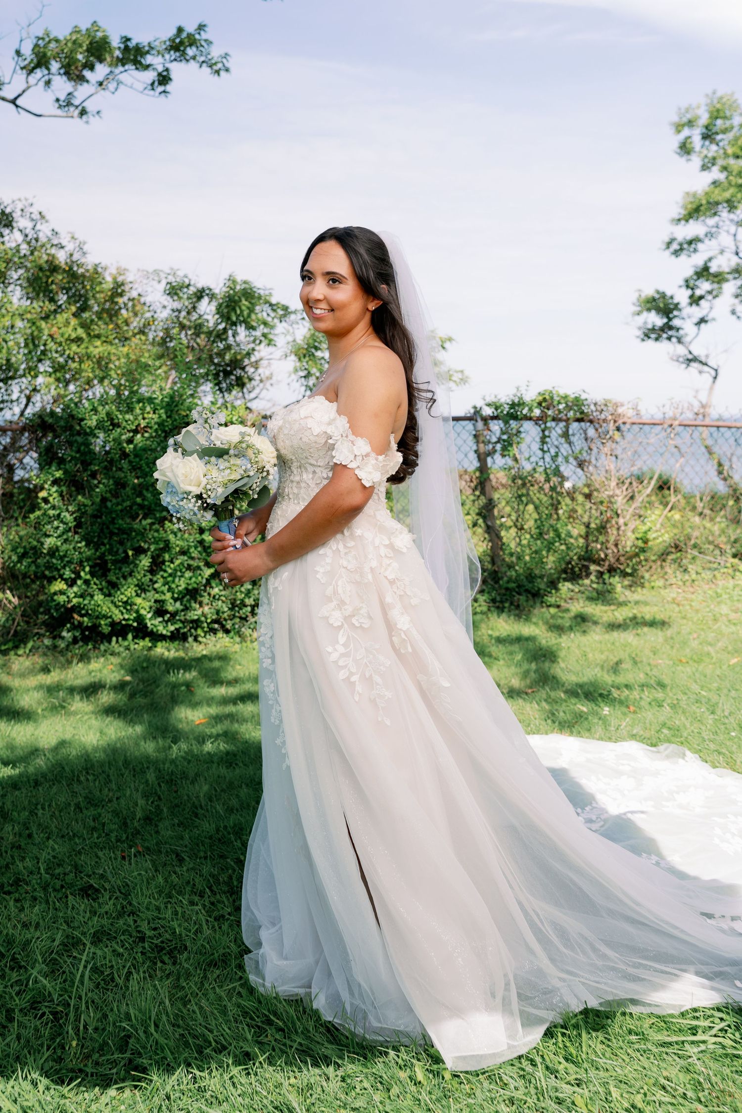 Bride in an off-shoulder lace wedding gown with a long veil and train holds a white floral bouquet on a sunny green lawn.