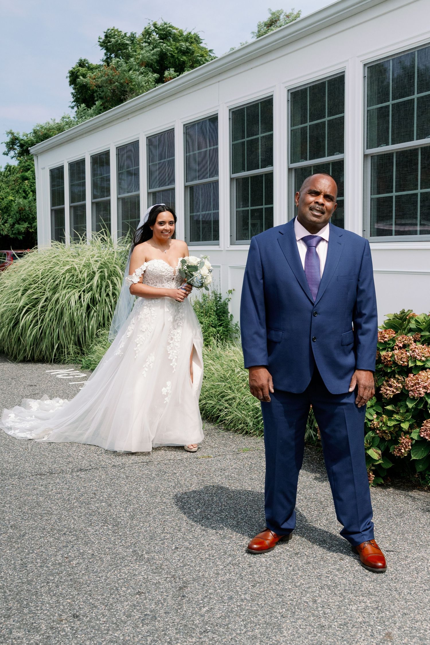 Bride in a white lace gown holding a bouquet walks toward a man in a blue suit and purple tie during a first look outdoors.