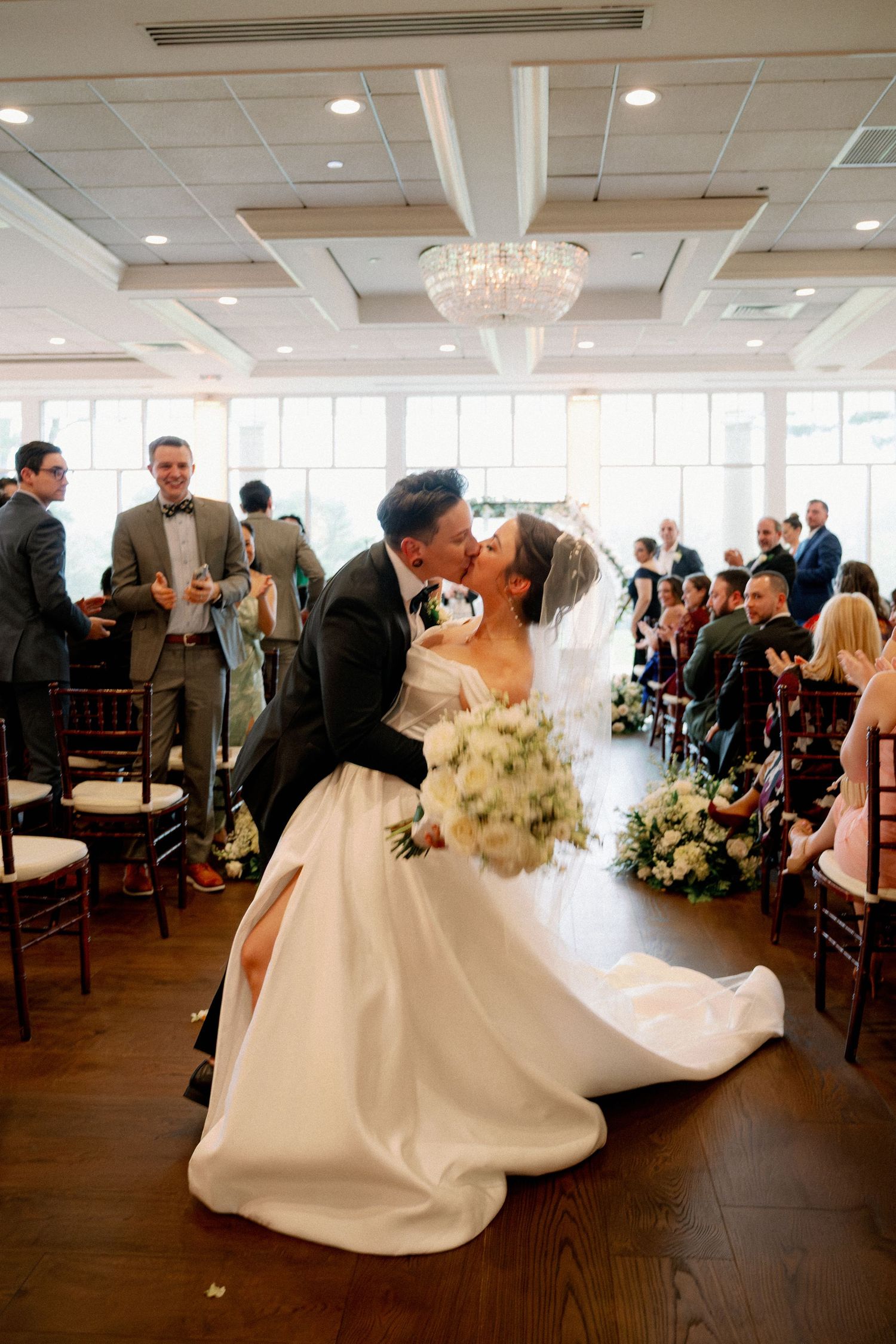 Bride and groom share a kiss in the aisle after their indoor wedding ceremony, guests applauding around them.