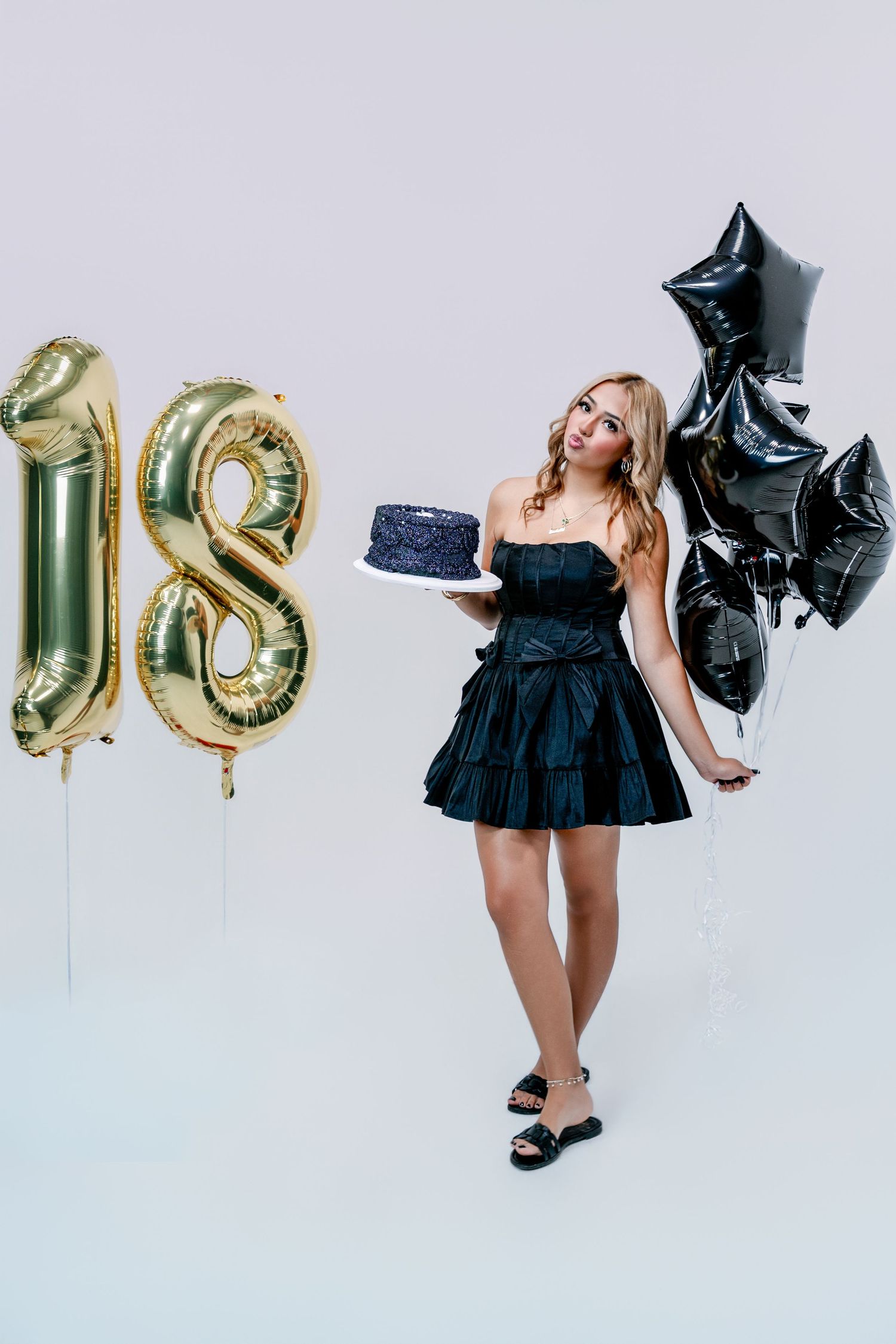 Young woman in a black ruffled dress holds a blue sparkly cake and black star balloons beside gold number 18 balloons for her birthday.