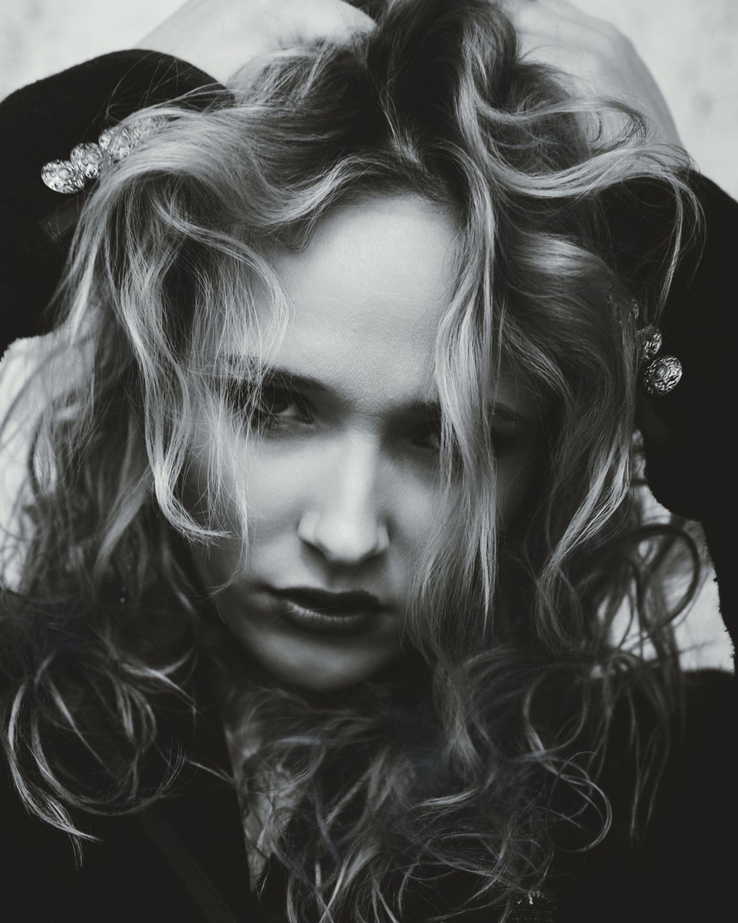 Black and white portrait of a woman with tousled wavy hair, hands raised to her head, wearing sparkling hair clips and earrings.