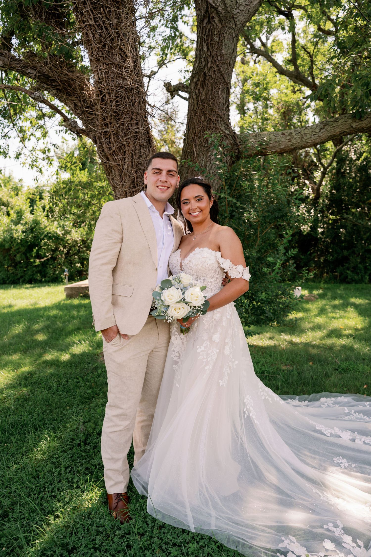 Smiling bride in a white lace gown holding a bouquet stands beside a groom in a cream suit under large trees at an outdoor wedding.