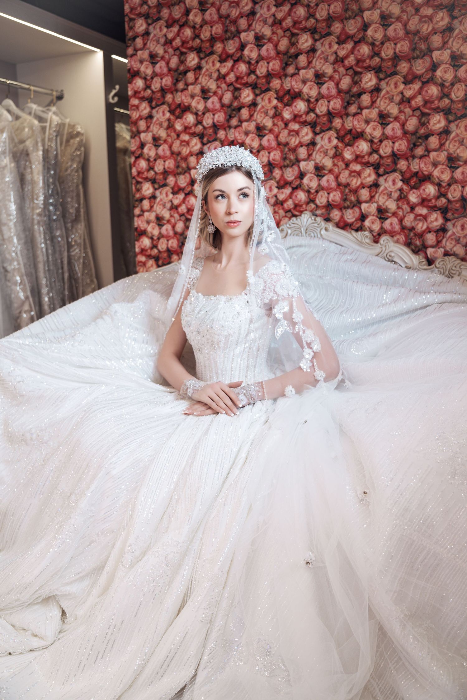 Elegant white wedding gown with lace veil against pink floral backdrop in bridal boutique.