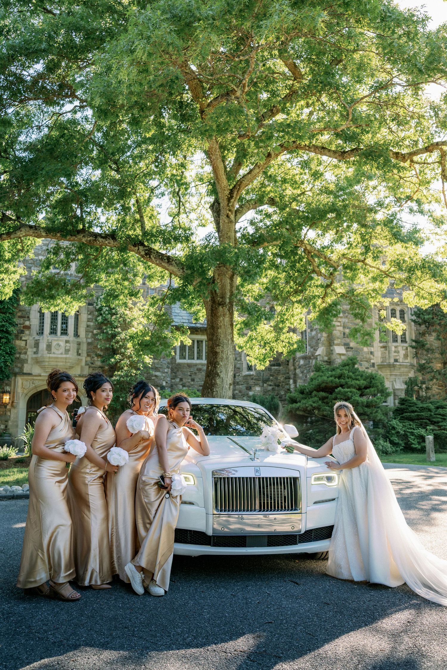 Bride in white gown poses with bridesmaids in gold dresses beside a white Rolls-Royce at an elegant estate wedding.