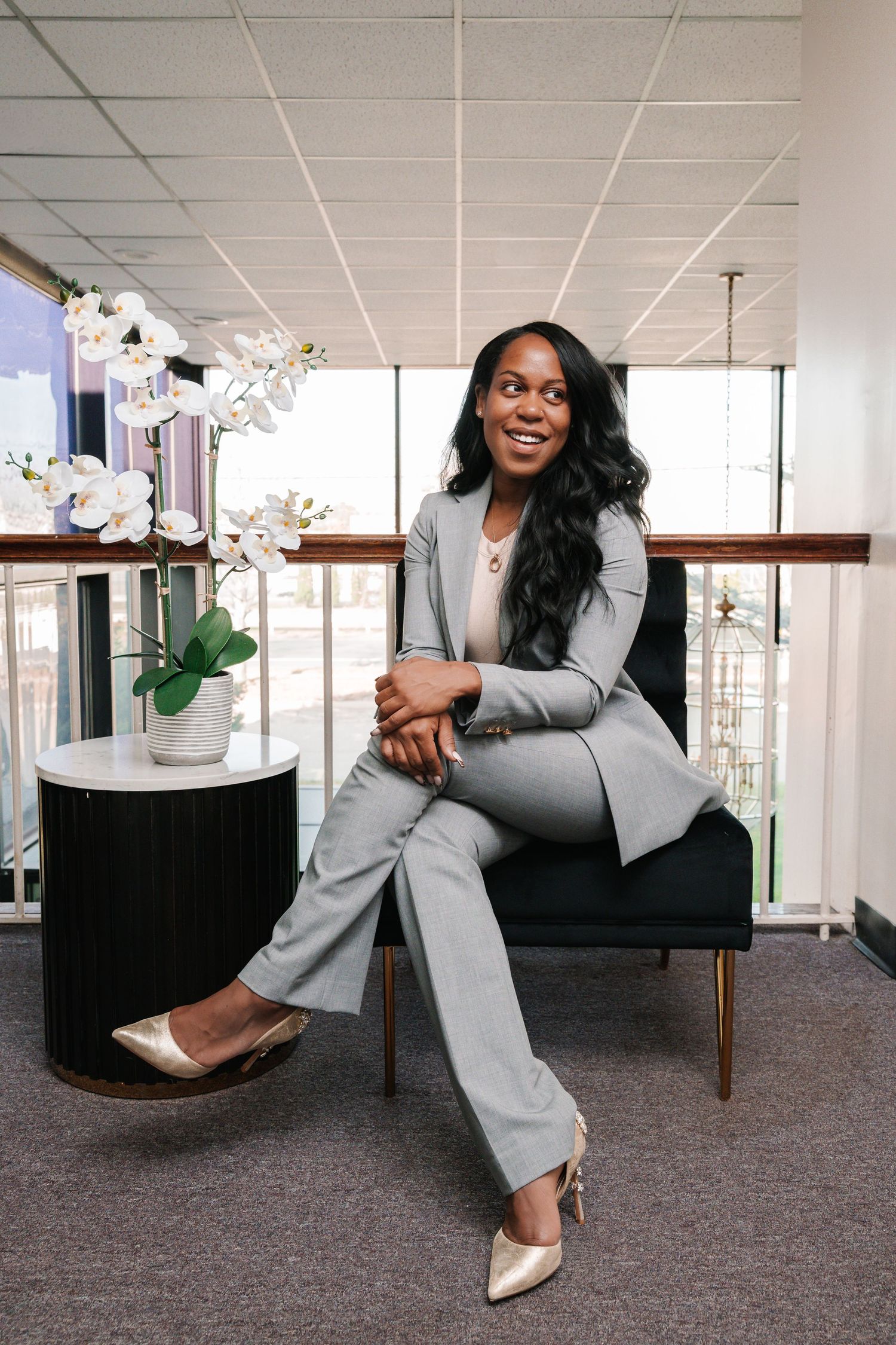 Smiling woman in a gray pantsuit and metallic heels sits on a black chair beside a white orchid in a bright modern office.