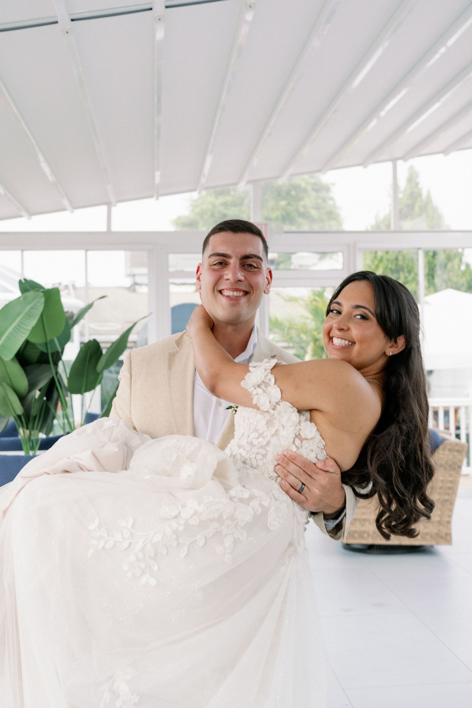 Smiling groom in a tan suit cradles his bride in a lace wedding gown inside a bright, airy sunroom with green plants.
