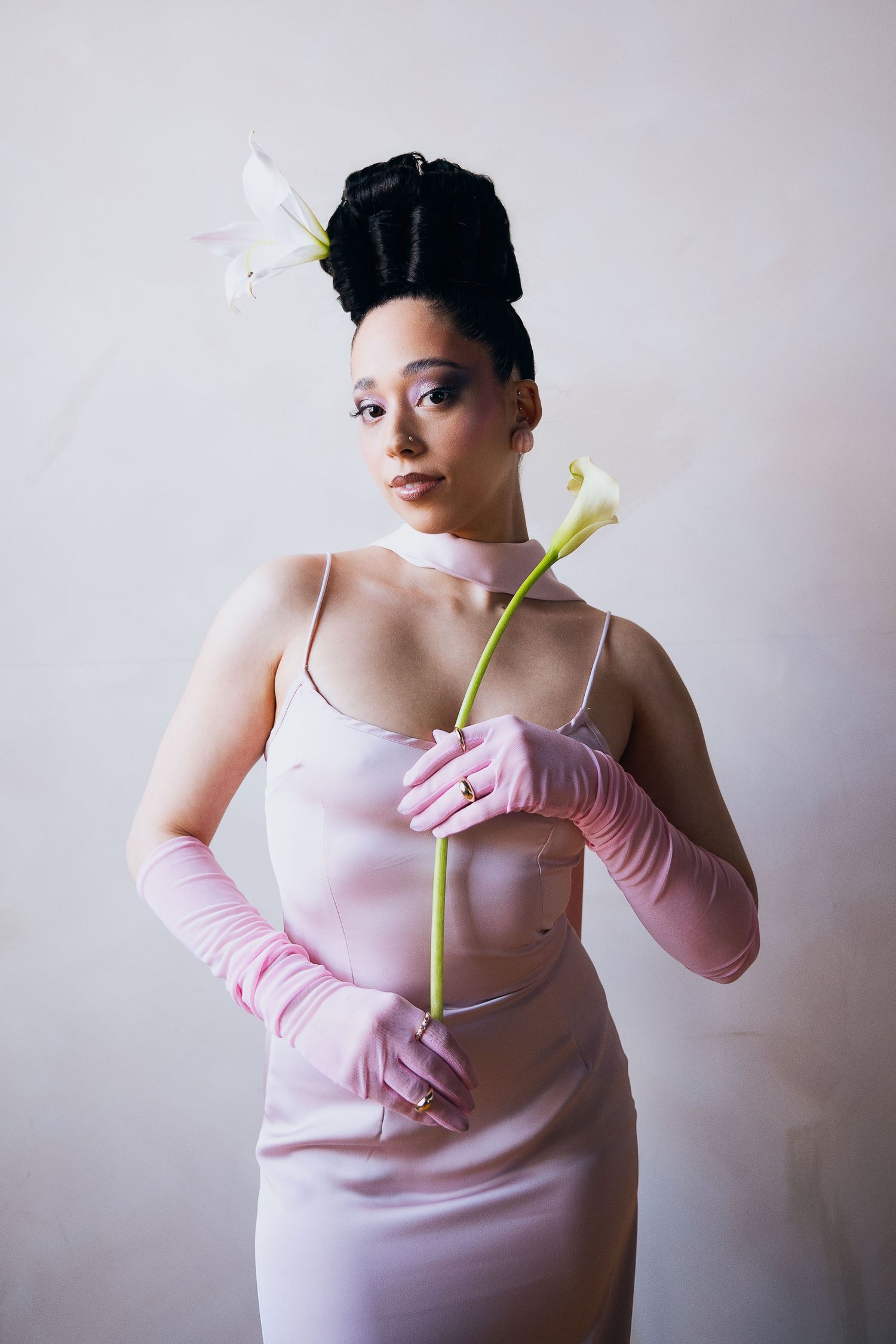 Woman in pink satin dress and gloves holds calla lily, with flowers in upswept black hair, against white background.
