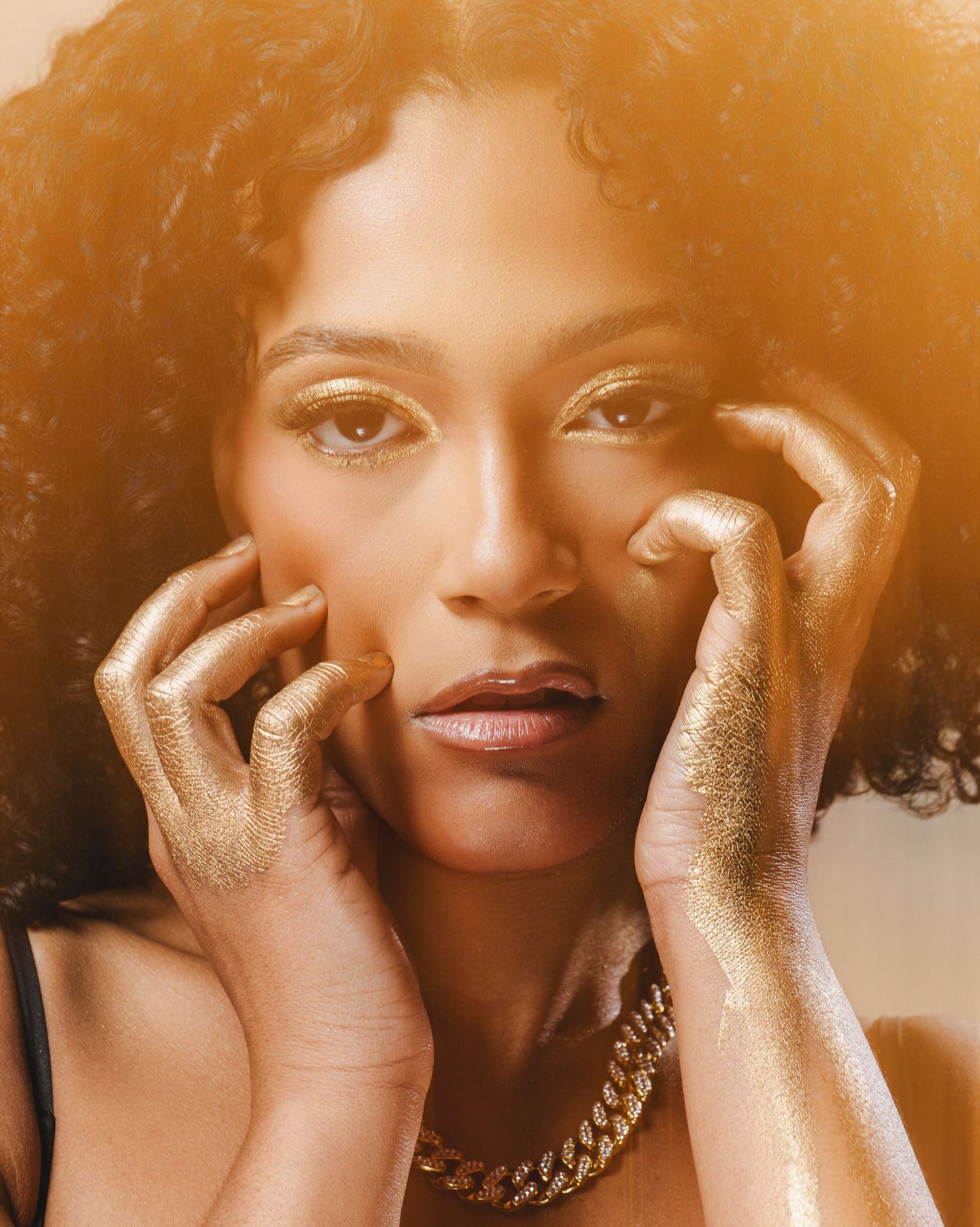 Portrait with golden glitter makeup and curly hair against warm lighting wearing a gold chain necklace.