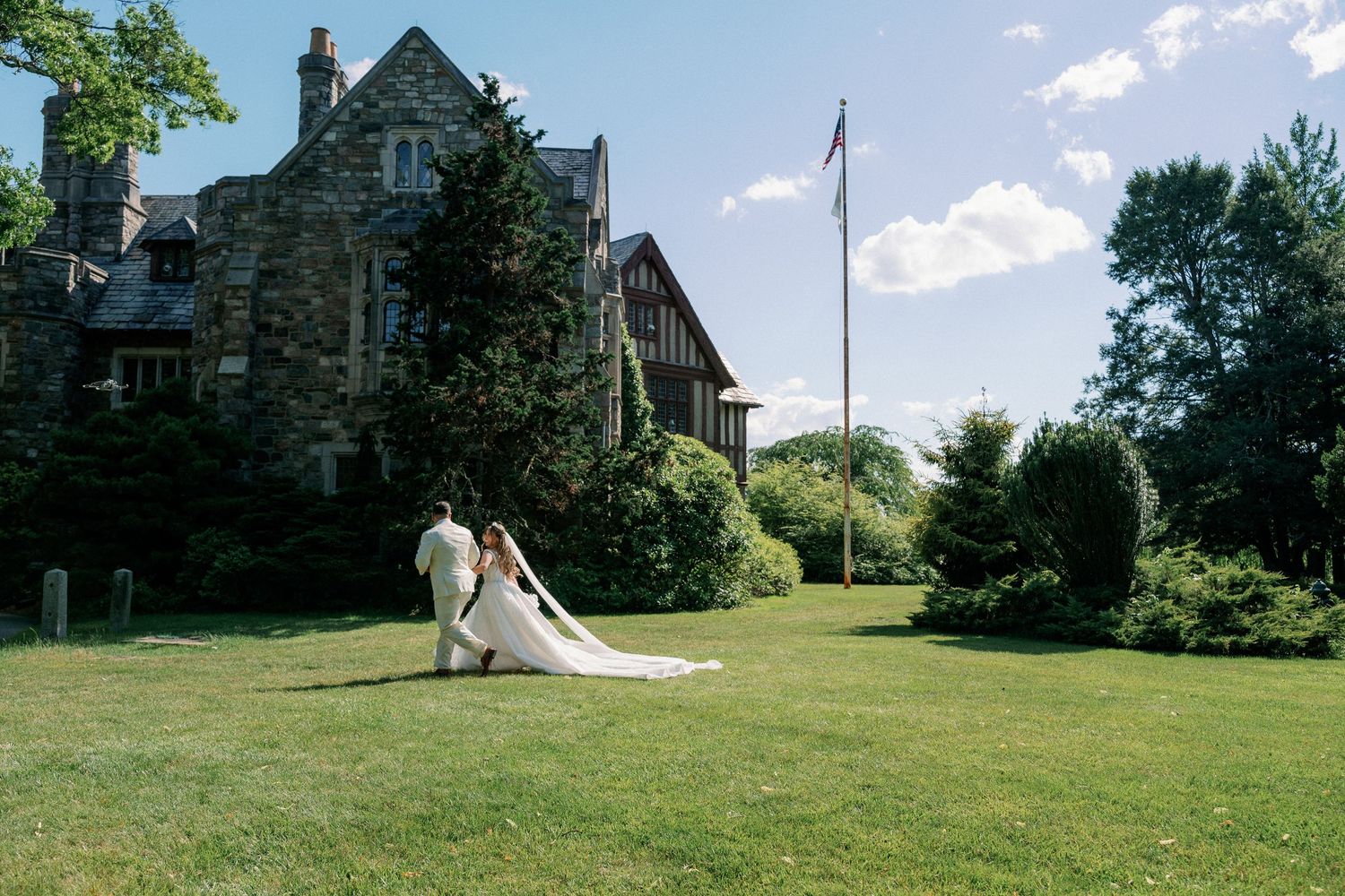 Bride in flowing white gown poses on lush green lawn before a grand stone manor house on a sunny day.