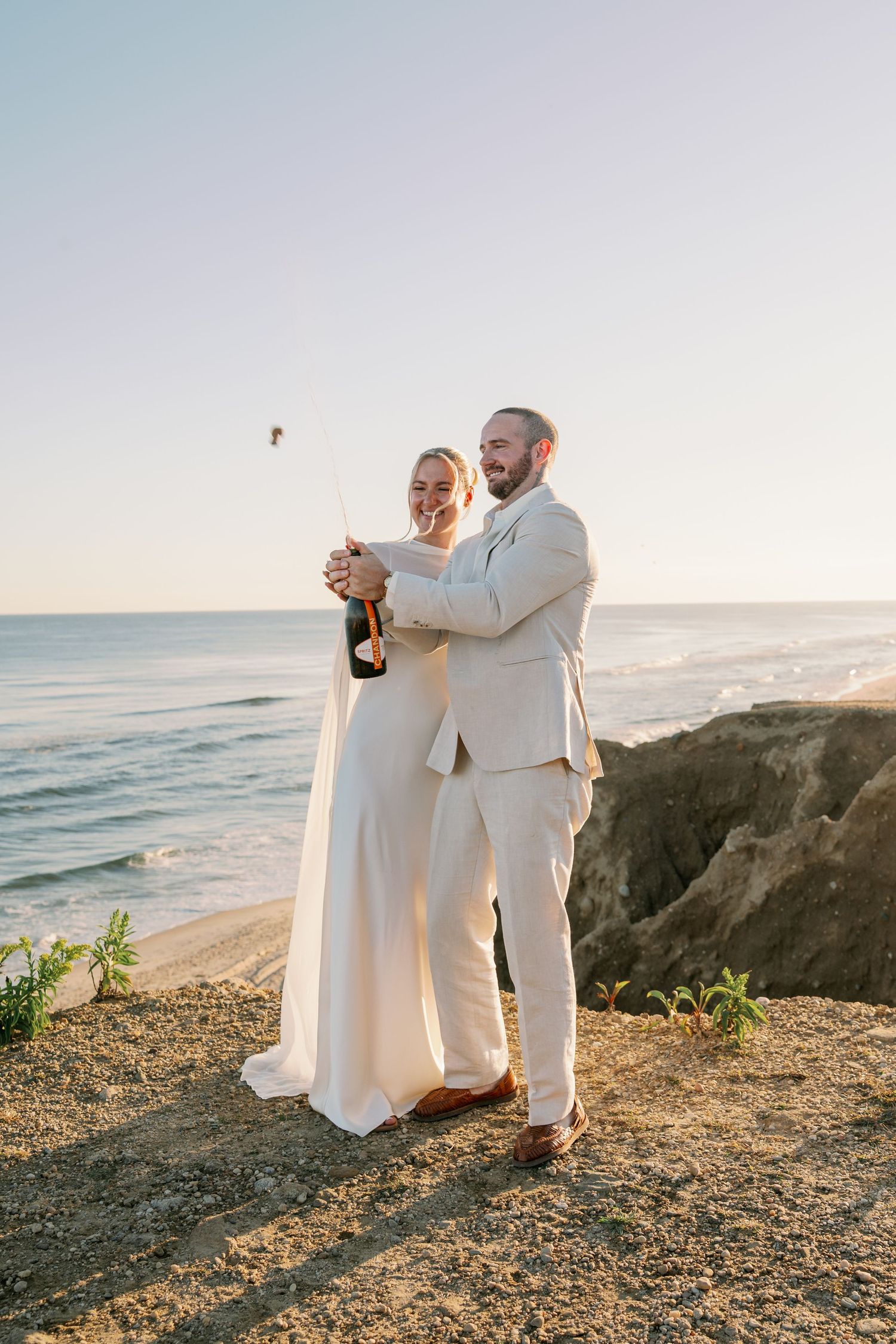 Bride in a white gown and groom in a cream suit pop champagne on a sunny coastal bluff overlooking the ocean at sunset.