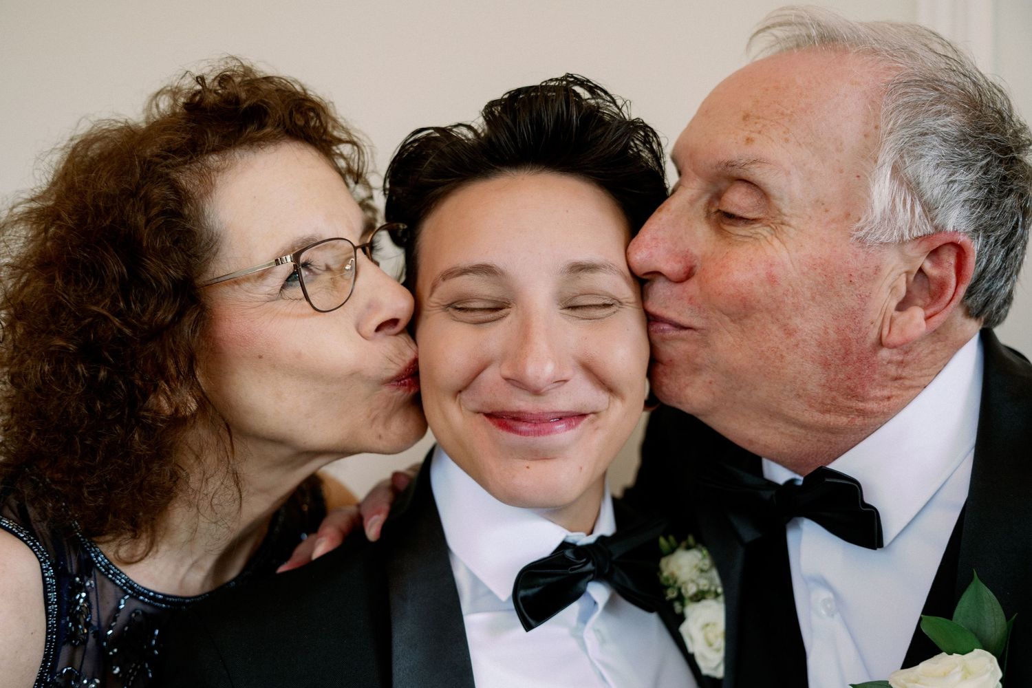 A young person in a tuxedo smiles as two older adults in formal wear kiss them on each cheek at a wedding.