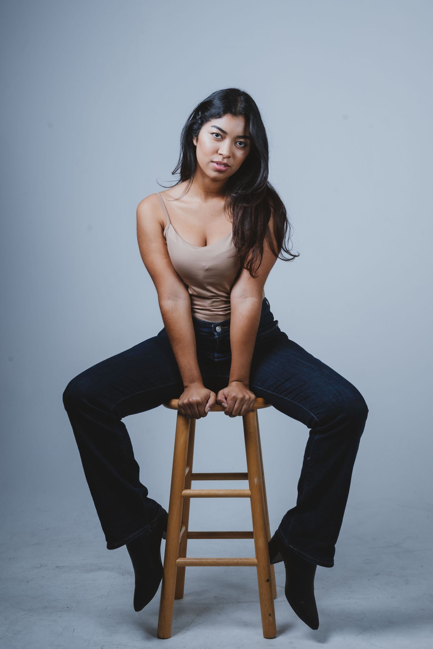 A model in a beige tank top and dark jeans poses casually on a wooden stool against a gray studio backdrop.
