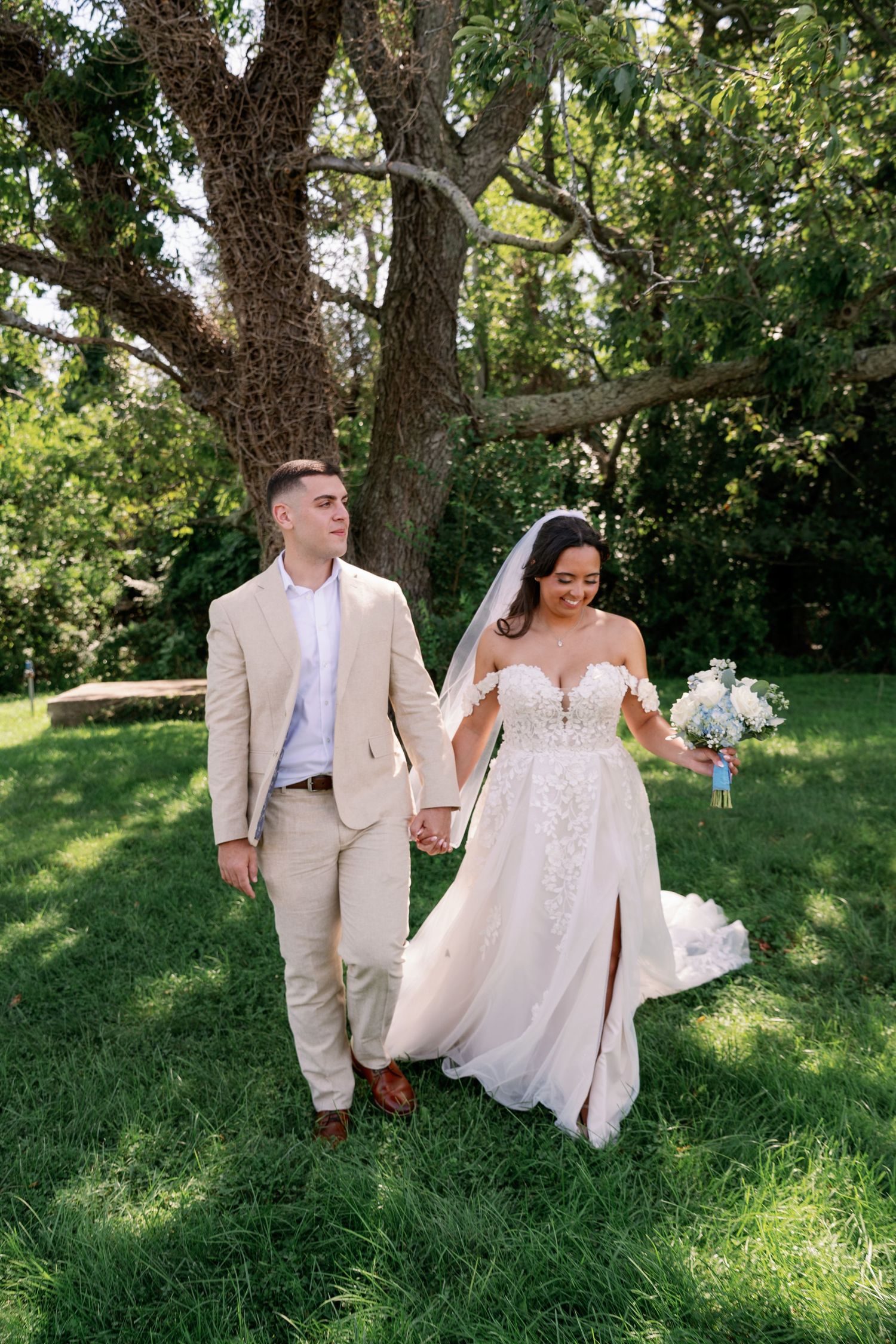 Bride in a lace off-shoulder gown and groom in a tan suit hold hands while walking across a sunny grassy lawn beneath large oak trees.