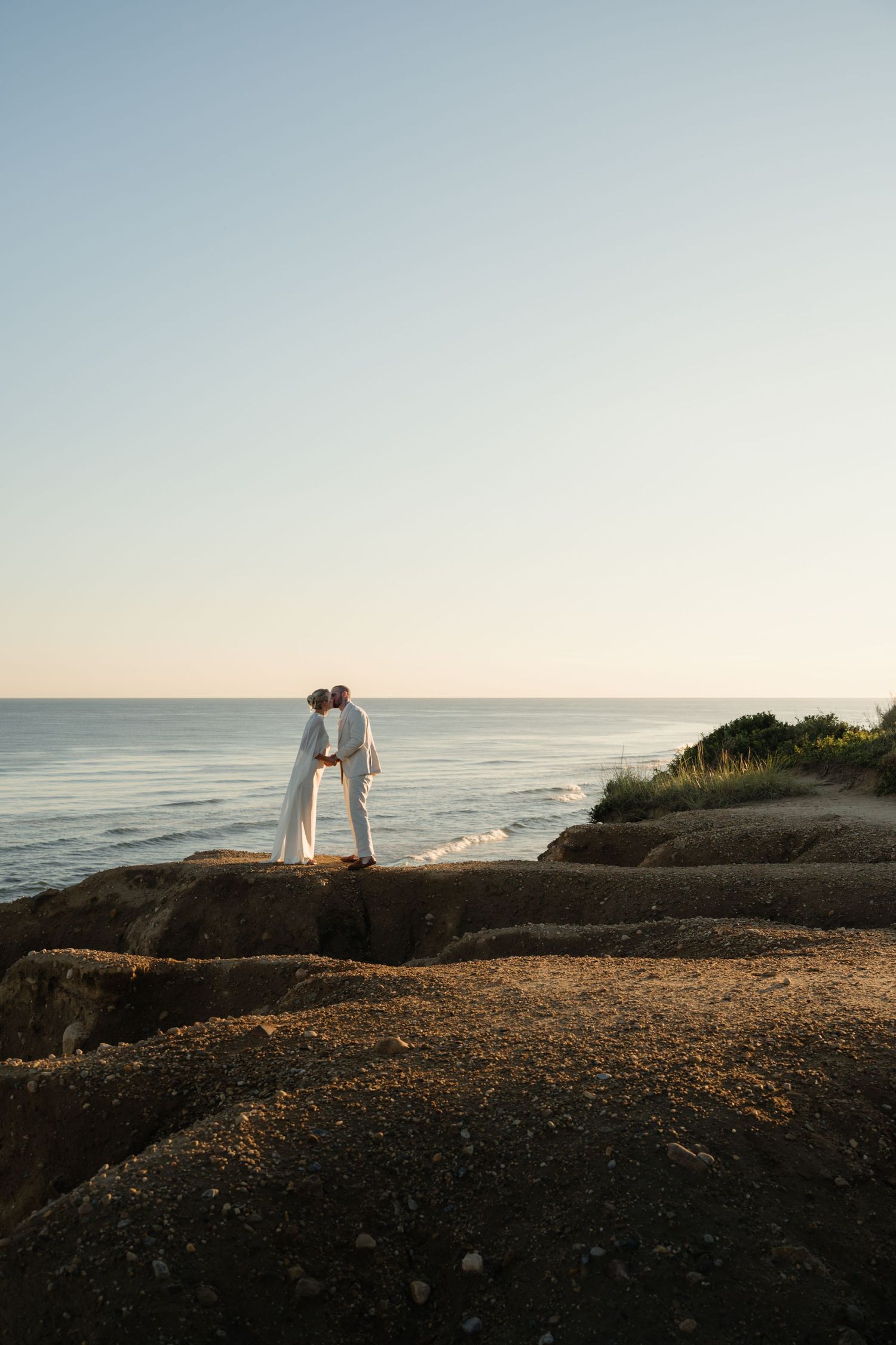 Couple in white embracing on a sandy coastal cliff edge at sunset, overlooking the calm ocean and clear horizon.
