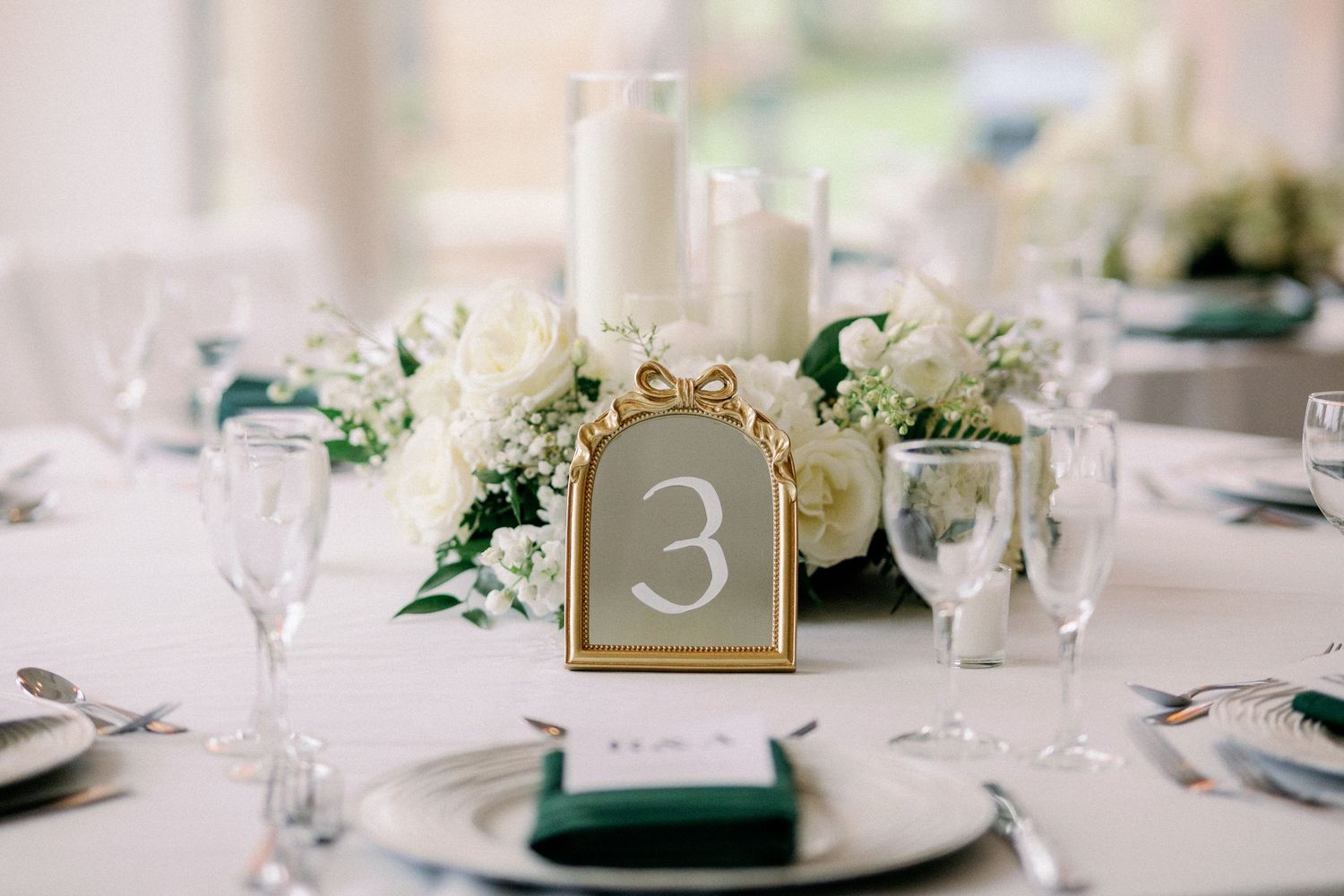 Elegant wedding table setting with white floral centerpiece, gold-framed table number 3, green napkins, and white candles.