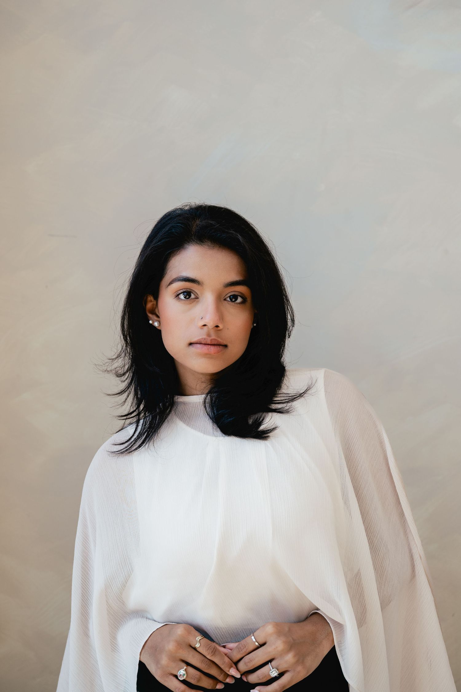 Woman with shoulder-length black hair in a flowing white cape blouse poses with hands clasped, wearing rings, against a soft neutral wall.