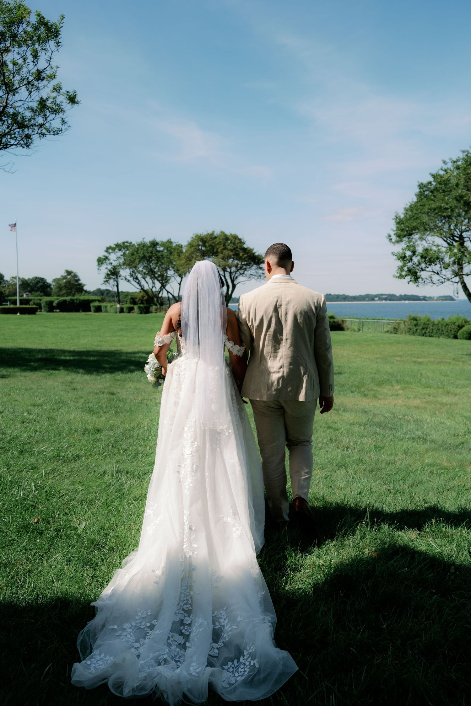 Bride in a lace gown with long veil and groom in a tan suit walk hand in hand across a sunny green lawn toward the waterfront.