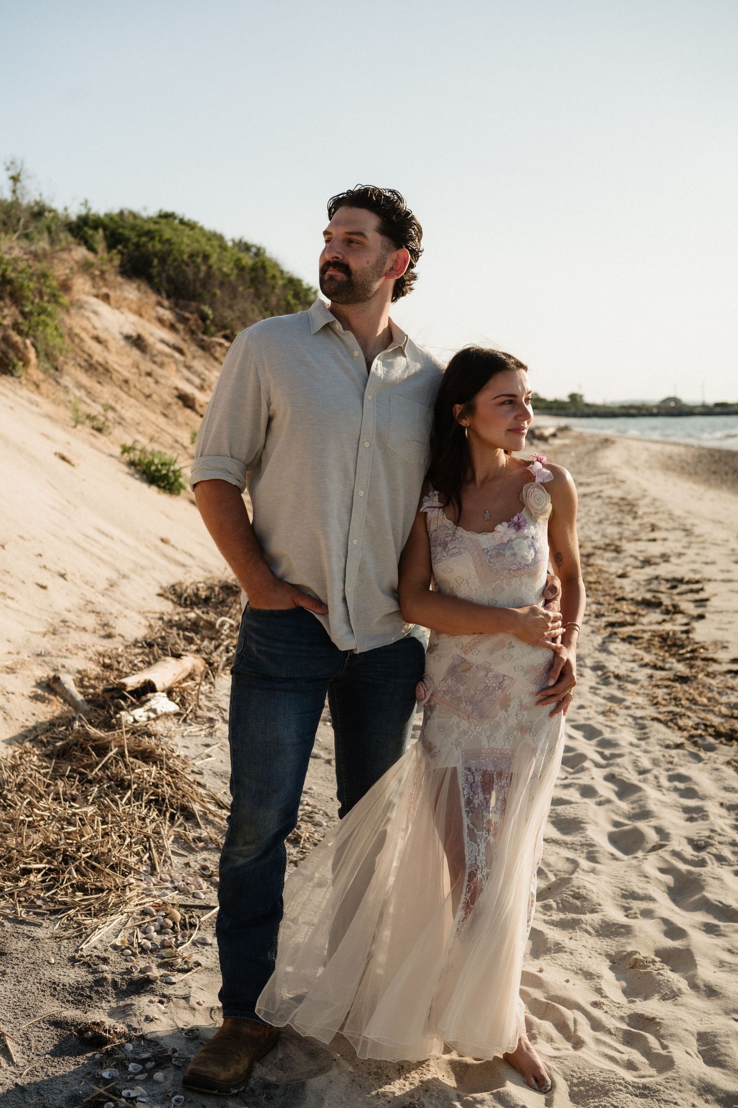 Couple embracing on a sandy beach at golden hour, woman in floral dress, man in jeans and linen shirt.