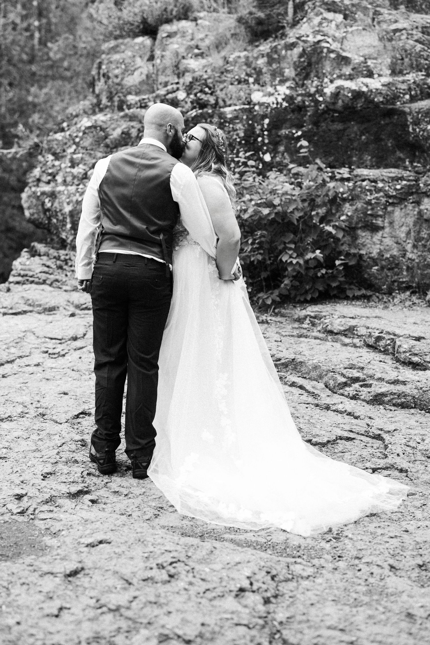 A wedding couple stands together on rocky terrain in black and white.