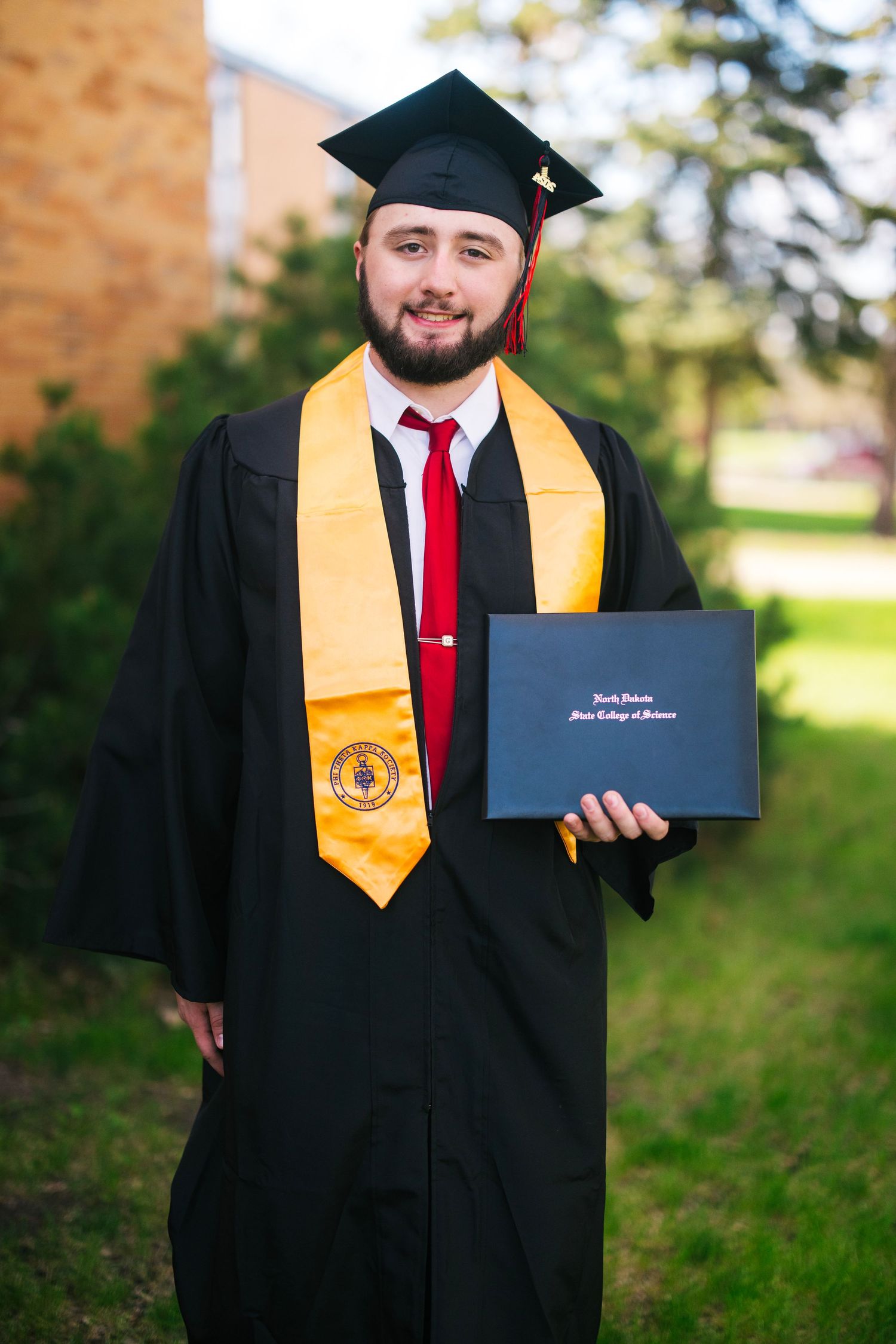 Graduate in cap and gown holding diploma with gold honors stole.