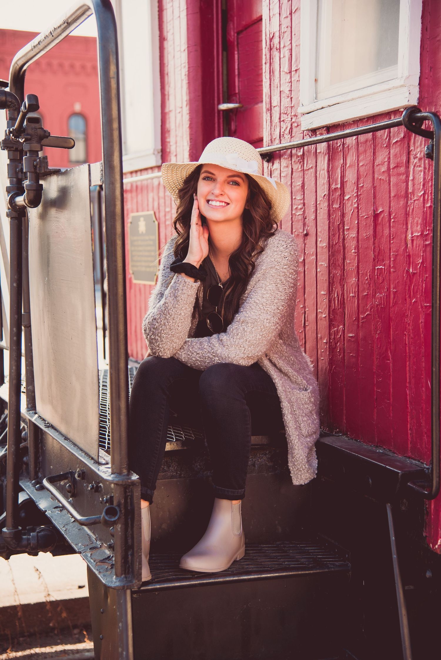 A person in a gray sweater and black pants poses playfully against a red brick wall with metal railings.