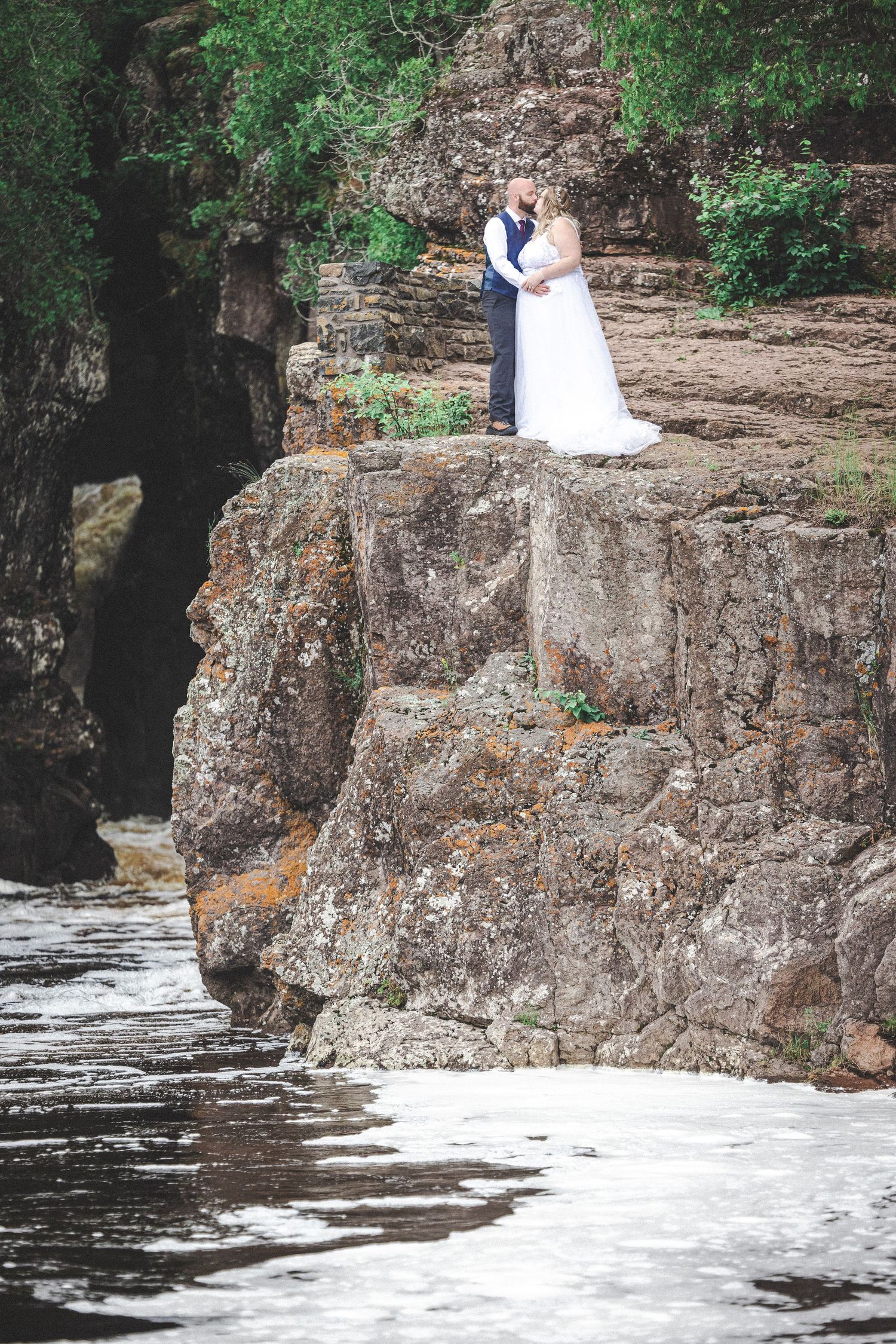 A wedding couple stands on dramatic cliffs overlooking rushing waters below.