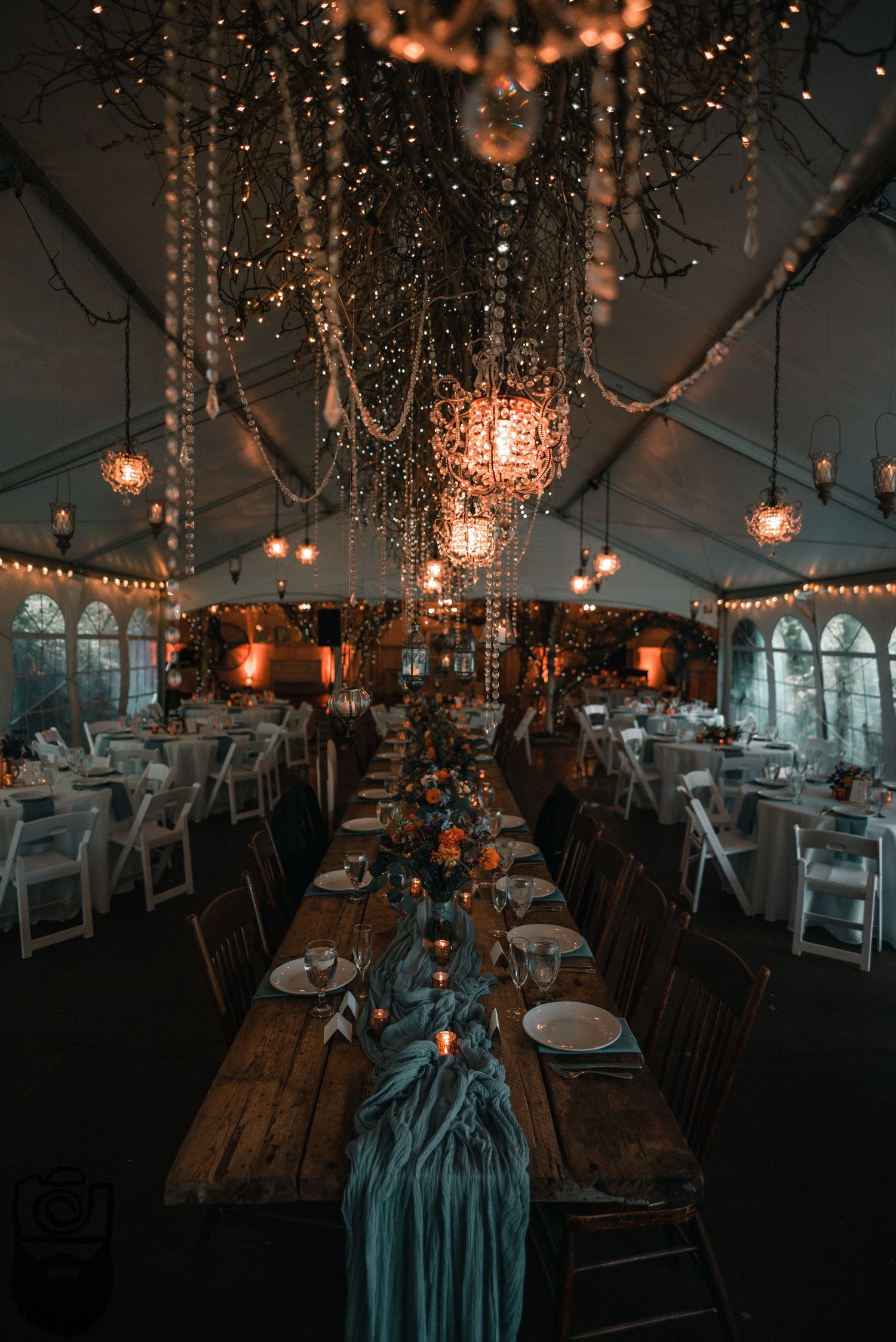 Elegant wedding reception tent decorated with hanging lights and crystal chandeliers over rustic wooden tables.